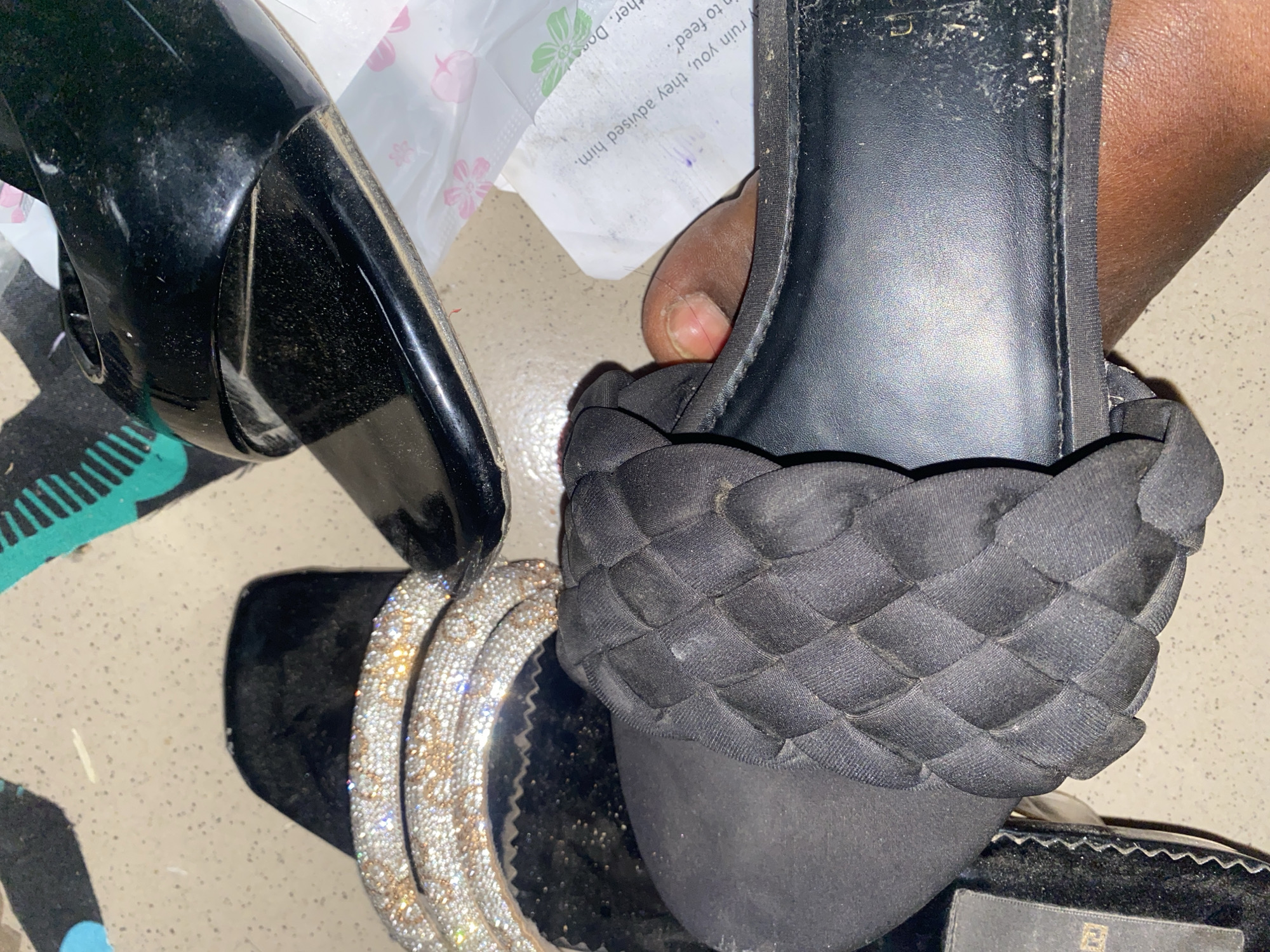 A person's feet are wearing two different black sandals on a speckled floor. One sandal has a strap adorned with rhinestones and gold-toned accents. The other sandal features a woven black strap and a smooth black sole. A piece of paper with printed text is visible beneath the footwear. The scene appears to be indoors, possibly a residential area in Jauro Yinu, Nigeria, given the context provided.
