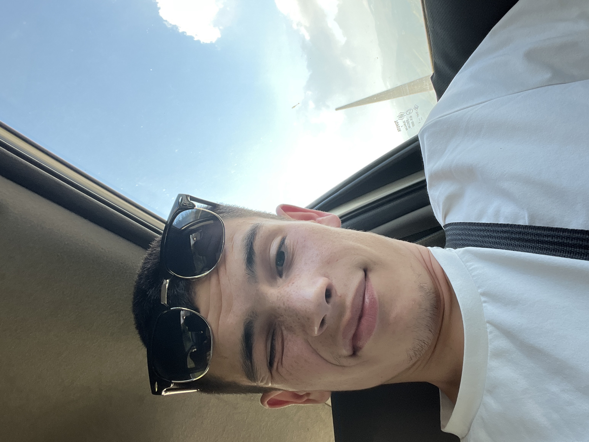 A young man is taking a selfie in a car, looking up towards the open sunroof. He is wearing sunglasses pushed up on his forehead and a white t-shirt, with a seatbelt visible across his chest. The sky is visible through the sunroof. There is no indication of the location being Tazha, Bulgaria.