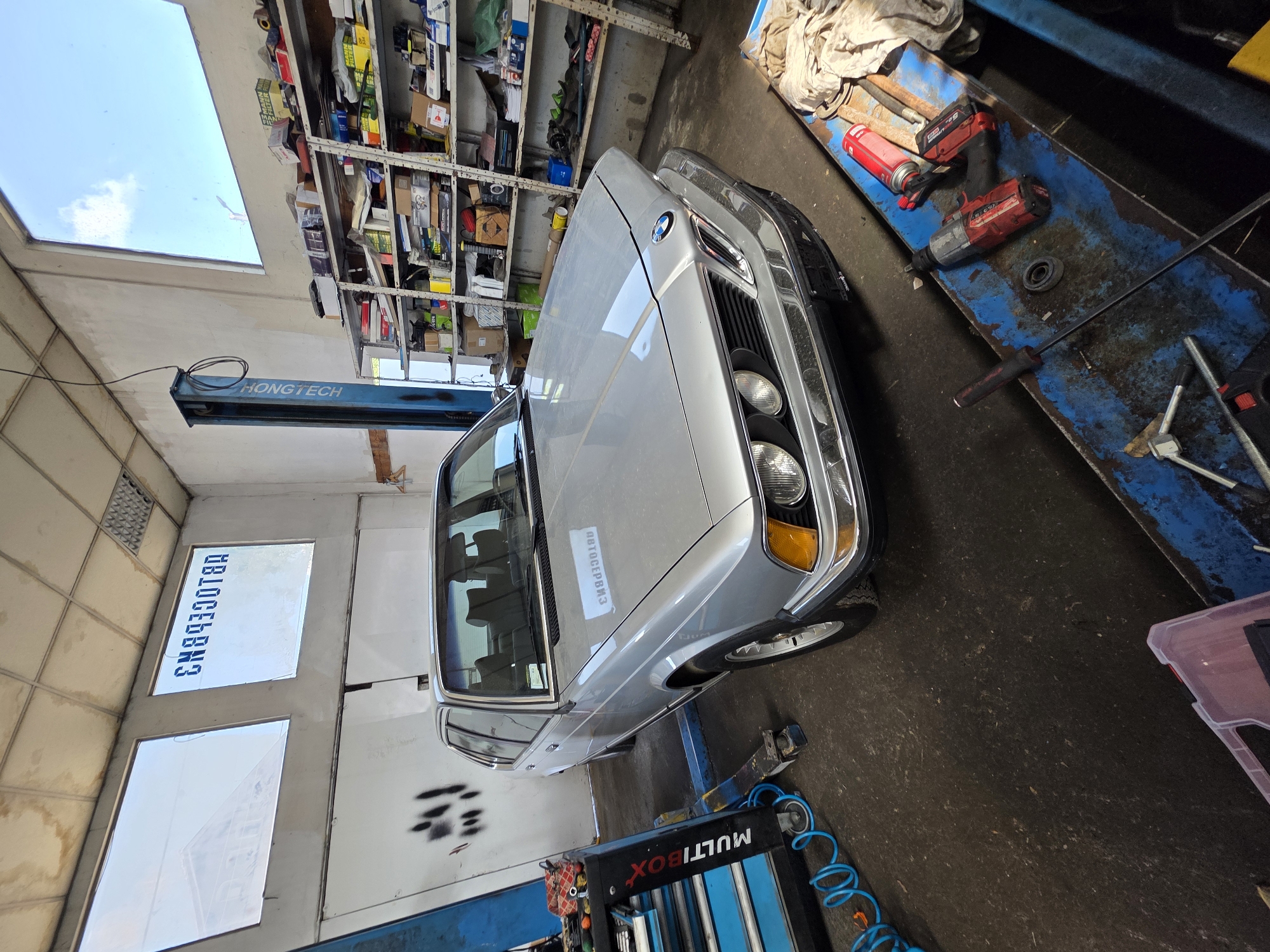 A silver BMW E30 is parked in a garage in Dragichevo, Bulgaria. The car is facing forward and is slightly angled to the left. The garage has a concrete floor and white walls. There are two windows in the back wall, one with a reflection of a building and the other with blue lettering that reads "CN8930T8A". A blue car lift is positioned to the left of the car. Shelves stocked with car parts are visible on the right wall. A tool cart with various tools is to the left of the car. A workbench with tools and car supplies is in the foreground. The overall scene suggests a car repair or maintenance shop.