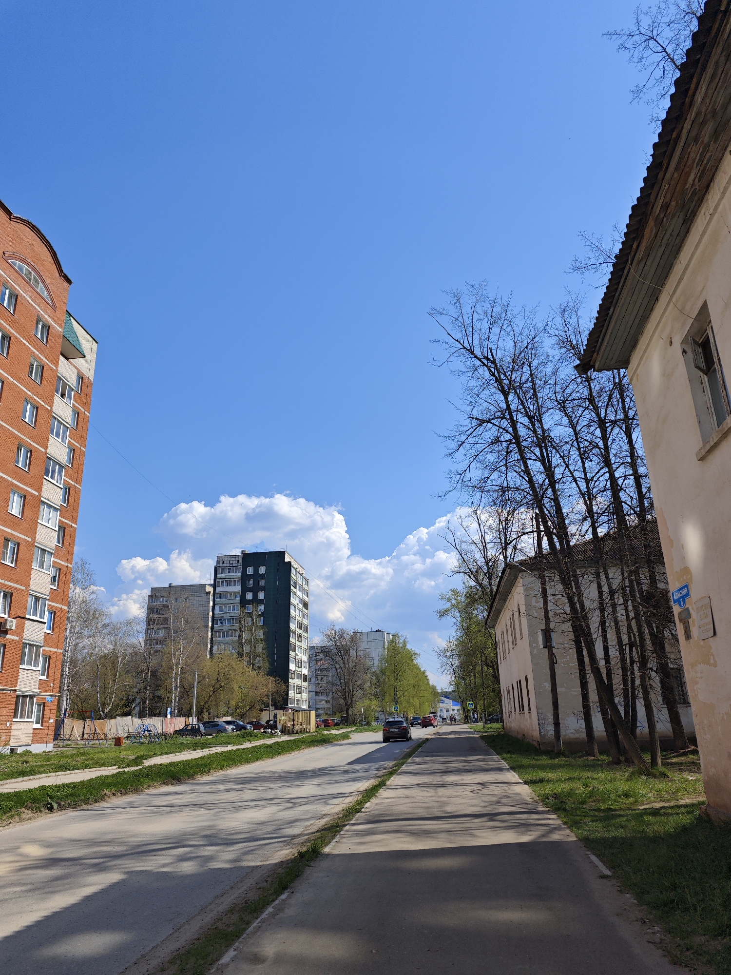 The image depicts a street scene in Chaikovsky, Russia. A paved road with a pedestrian sidewalk runs through the center of the frame. A red brick apartment building stands on the left, while an older, two-story building with a decaying facade is on the right. A row of bare trees lines the sidewalk on the right. In the distance, apartment buildings are visible against a blue sky with scattered clouds. A few cars are on the road. The scene is bathed in sunlight, casting shadows across the road and sidewalk.