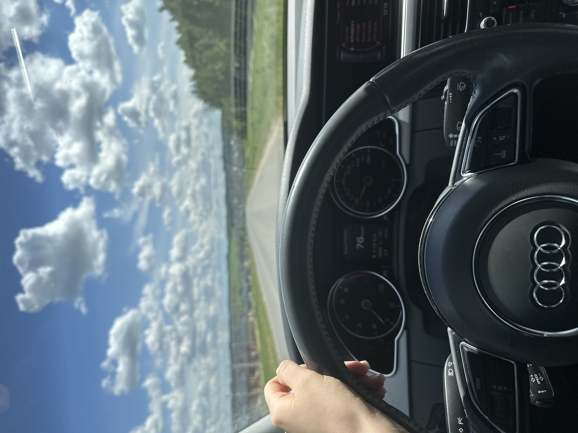The image shows the interior of an Audi car, specifically the steering wheel and dashboard, as seen from the driver's perspective. The steering wheel is prominently displayed, featuring the Audi logo in the center. Part of the dashboard is visible behind the steering wheel, along with the instrument panel displaying a speed of 76. A hand is holding the bottom of the steering wheel.
The scene outside the car is visible through the windshield, showing a road stretching into the distance, flanked by green fields and trees. A cloudy blue sky dominates the upper portion of the view. The scene appears to be a rural or suburban area. Эстачи, Russia is not identifiable in the image.