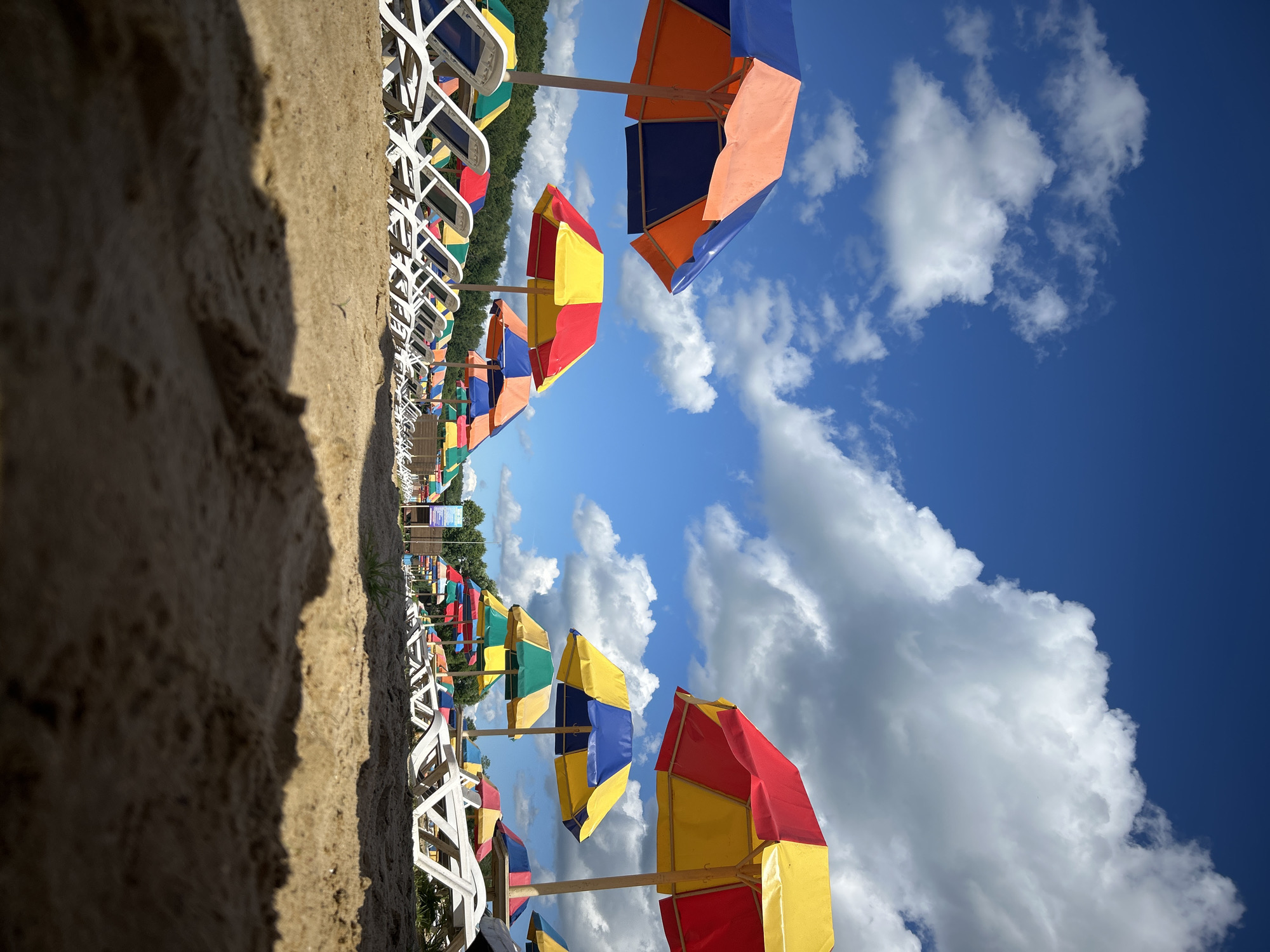 The image depicts a sunny day at a beach or park with rows of empty lounge chairs and colorful umbrellas. The foreground is dominated by a sandy surface, with the camera positioned at a low angle, capturing the texture of the sand. In the midground, multiple rows of white lounge chairs are visible, each partially shaded by multi-colored umbrellas. The umbrellas are predominantly red, yellow, blue, and orange, arranged in a pattern. Beyond the chairs and umbrellas, a line of green trees forms the background, suggesting a natural setting. The sky is a vibrant blue with scattered white cumulus clouds, indicating clear weather. The scene is static and does not show any people or immediate activity. The location is identified as the city of Юбилейный, Russia.
