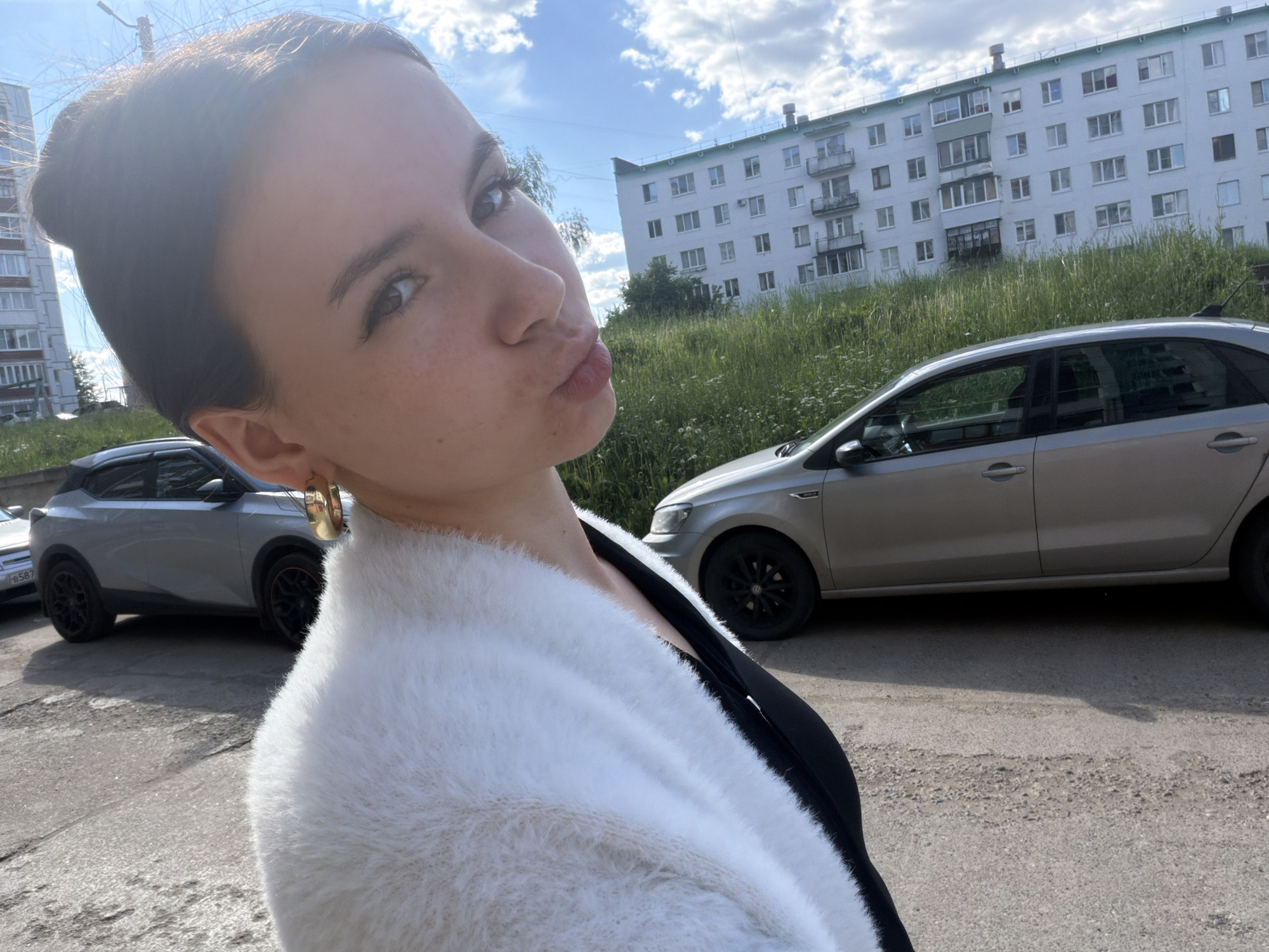 The image depicts an outdoor daytime scene in a residential area of Chaikovsky, Russia.
The primary subject is a young woman, captured in a close-up selfie-style shot, angled from her right side. She has dark hair pulled back, a fair complexion with visible freckles across her nose and cheeks. Her dark eyes, accentuated with eyeliner, gaze directly at the camera. Her lips are pursed in a "kiss" gesture, coated with a light pink or reddish hue. A gold-colored hoop earring is visible in her left ear. She is wearing a white, fluffy or fuzzy cardigan over a dark, likely black, top or dress.
In the background, under a bright blue sky with scattered white cumulus clouds, stands a multi-story apartment building. This structure is light-colored, possibly off-white or pale blue, with numerous rectangular windows and several enclosed and open balconies. A distinctive light green coping or trim runs along its roofline. A second, similar apartment building is partially visible on the far left.
Two cars are parked on the asphalt surface in the mid-ground. On the right, a silver or light grey five-door hatchback (resembling a Volkswagen Polo) is visible, featuring dark-colored wheels. On the left, a grey compact SUV (possibly a Nissan Juke) is parked, displaying dark wheels with subtle red or orange trim accents. A partial Russian license plate, "B 587," is discernible on the front of the SUV.
Between the parking area and the apartment buildings, a strip of tall, overgrown green grass and weeds with small white flowers separates the street from the structures. The ground beneath the vehicles is a worn asphalt surface with visible cracks and uneven patches. The scene is illuminated by strong sunlight, indicating a clear, sunny day.
