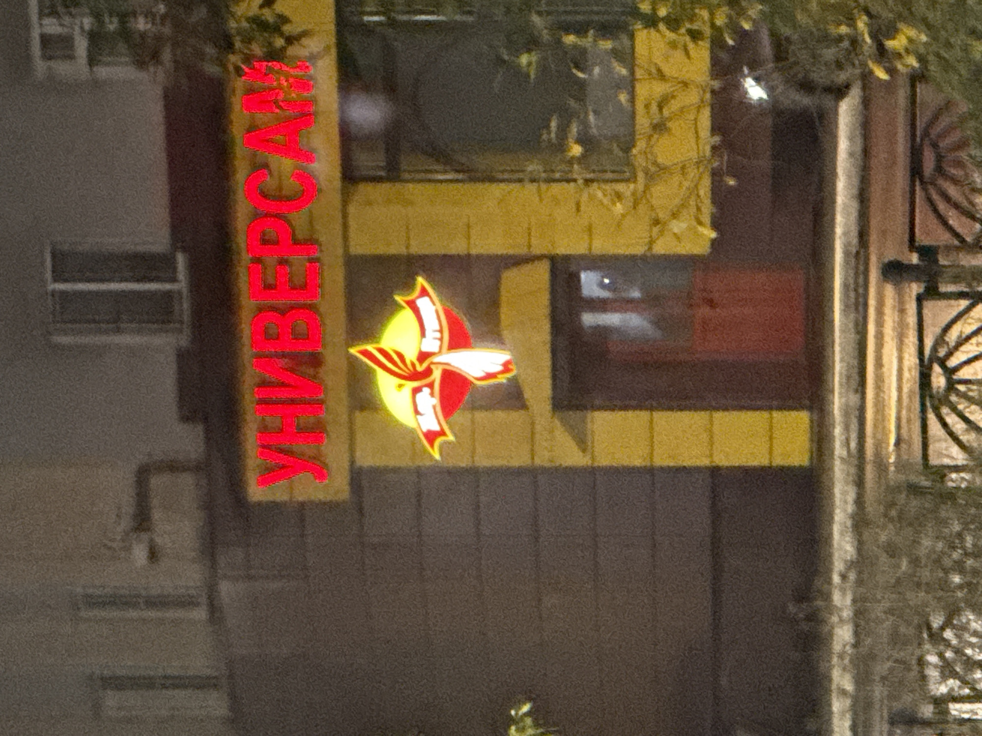 The image shows a neon sign that reads "УНИВЕРСАМ" (UNIVERSSAM) in red letters against a yellow background. Below the sign is a stylized logo of a bird with wings spread, also in red and yellow. The sign appears to be on the exterior of a building, likely a store. The building has dark gray brickwork and several windows. A red door is visible below the logo. The scene is illuminated by artificial light, suggesting it is nighttime or a dimly lit area. The foreground shows some foliage and a metal fence. The image is grainy, indicating it was taken with a low-light camera or at a distance. Based on the text on the sign and common store naming conventions in Russia, this is likely a "Universal Store" or supermarket. Without further information, the precise location in Norilsk, Russia cannot be determined, but it is a commercial establishment.