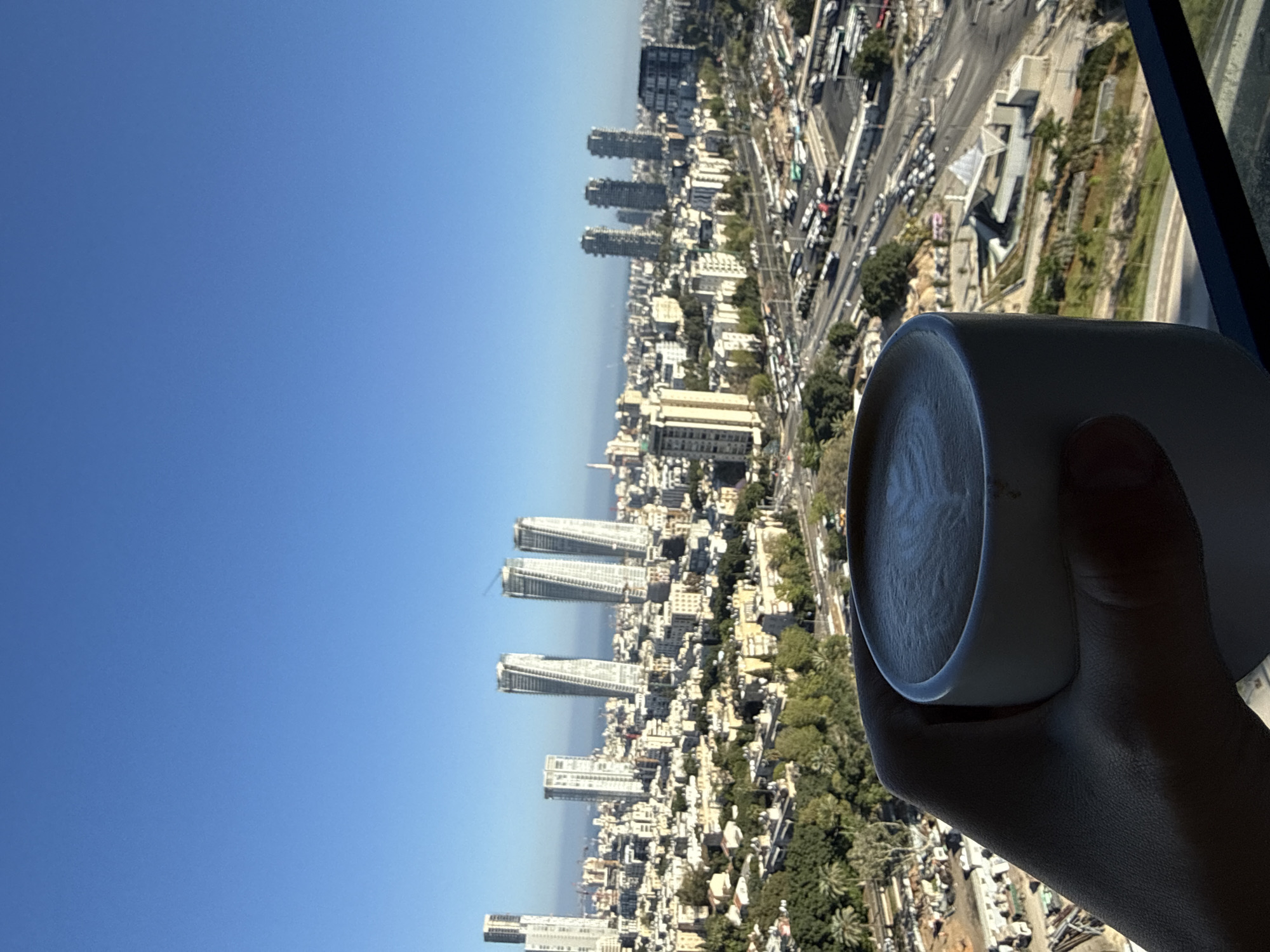 An arm and hand are holding a white mug, positioned in the lower right of the scene, against a backdrop of the city of Tel-Aviv, Israel, and a clear blue sky. The city features tall skyscrapers, residential buildings, and streets with visible car traffic, all bathed in bright sunlight. Vegetation is distributed throughout the city landscape. The mug appears to contain coffee.