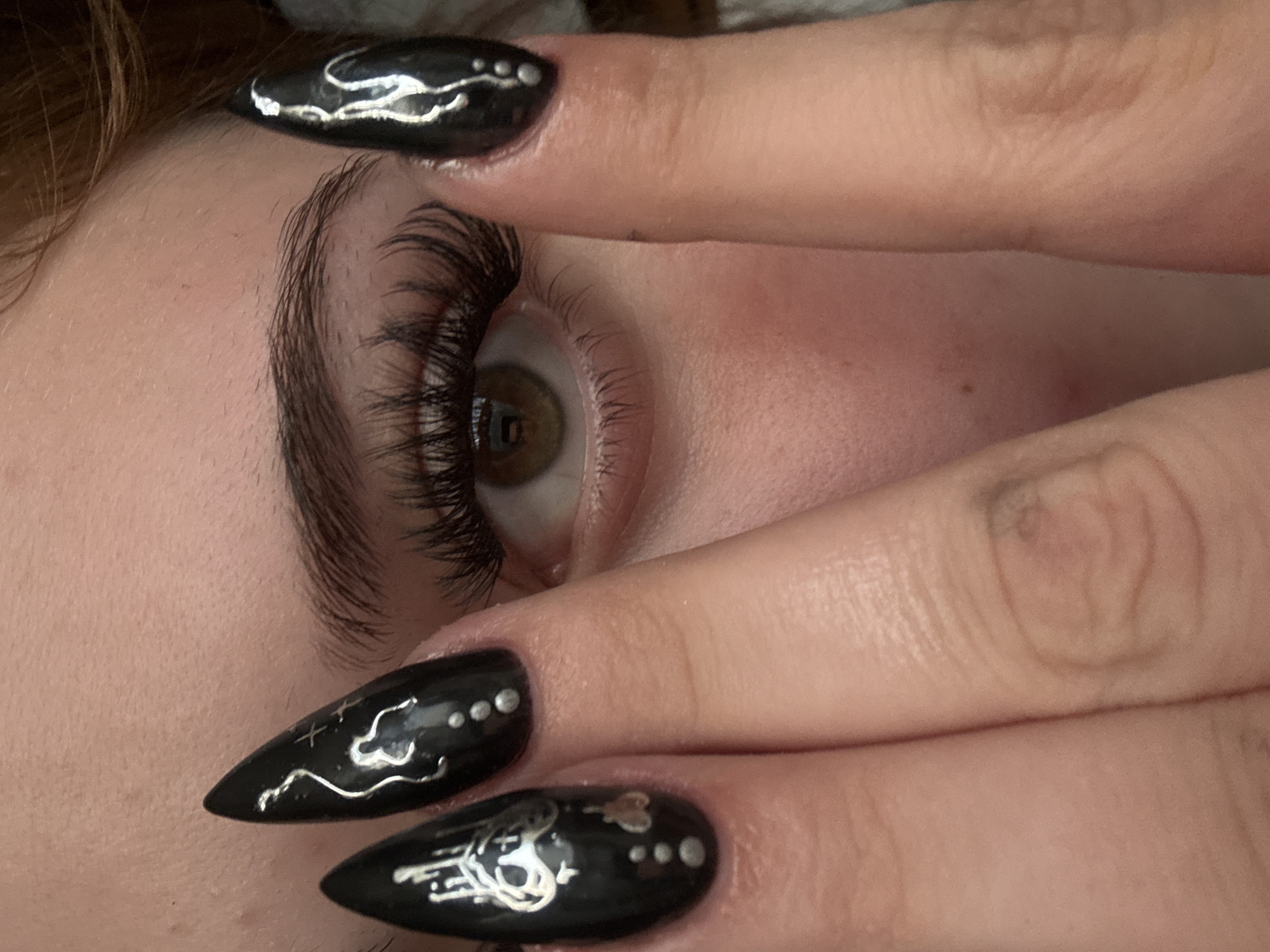 A close-up image displays the right eye of a person, mostly concealed by their hand. The visible eye features an olive-colored iris and long, artificial eyelashes. The person's eyebrow is dark and well-defined. Their fingers are adorned with long, black fingernails featuring silver designs, including dots and swirling lines. The skin is fair-toned, and no specific location in Troyan, Bulgaria, is discernible.