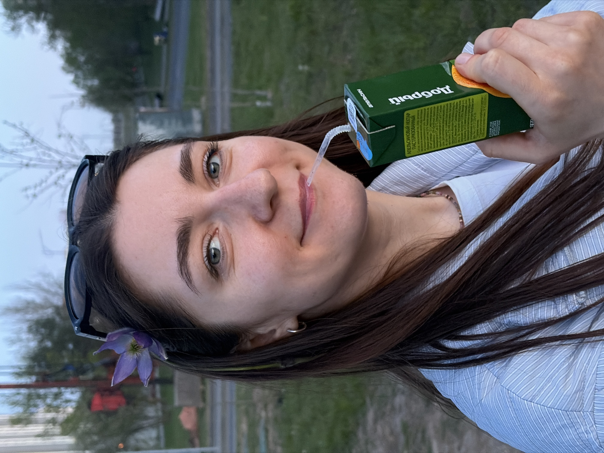 A young woman is pictured from the chest up, drinking from a green juice box with a straw. She is holding the juice box in her right hand. The woman has long dark hair, light skin, and green eyes. Sunglasses are placed on her head, and a purple flower is tucked behind her ear. She is wearing a light blue striped shirt. The background is a blurry urban scene, possibly a park or roadside, with greenery and a road visible. Given the "Добрый" brand of juice, which is popular in Eastern Europe, and the road and vegetation similar to Minsk, Belarus, it is possible that the location is Minsk, Belarus.