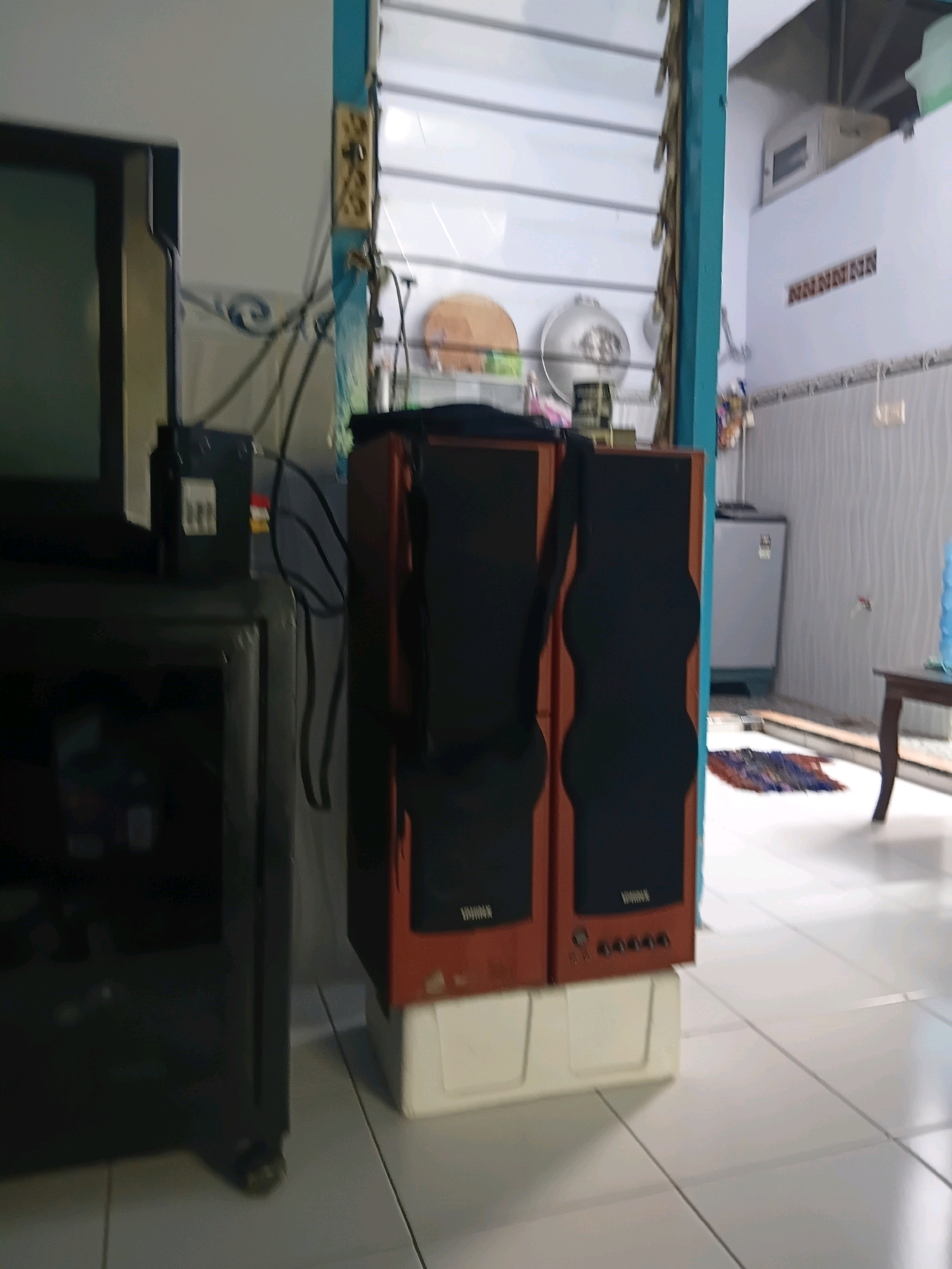 The image depicts an indoor scene, likely a room in Tanggul Wetan, Indonesia. In the foreground, a black electronic device, possibly a television or a computer monitor, is partially visible on the left. To its right, a pair of brown speakers with black fabric grilles are stacked on a white base. The speakers have visible branding and control buttons. Behind the speakers, a blue-painted vertical wooden beam is prominent, part of a structure that appears to be a staircase or a room divider. The background shows a light-colored wall with horizontal lines, suggestive of a tiled or paneled surface. Further in the background, a light gray refrigerator is visible, and a wooden table with a dark top is to the right. A woven mat is on the floor in front of the speakers. The overall impression is of a residential interior with common household items.