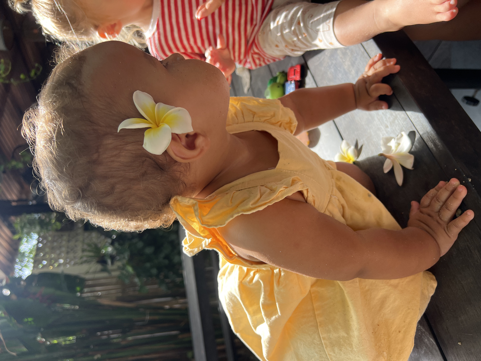 The image shows two young children on a dark wooden surface, likely a table, outdoors in North Kuta, Indonesia. The child in the foreground, a baby wearing a yellow dress, is looking upwards, with a white and yellow plumeria flower tucked behind their ear. Their arm is resting on the table, and their hand is visible. Another child, wearing a red and white striped shirt, is in the background, partially visible from the chest up. Sunlight is casting strong shadows, indicating it is likely daytime. The setting appears to be a casual outdoor area, possibly a restaurant patio or backyard, with some greenery visible in the blurred background. Two more plumeria flowers are visible on the table near the baby's hand, suggesting a tropical environment. The overall mood is peaceful and captures a candid moment of childhood. There is no visible text in the image.