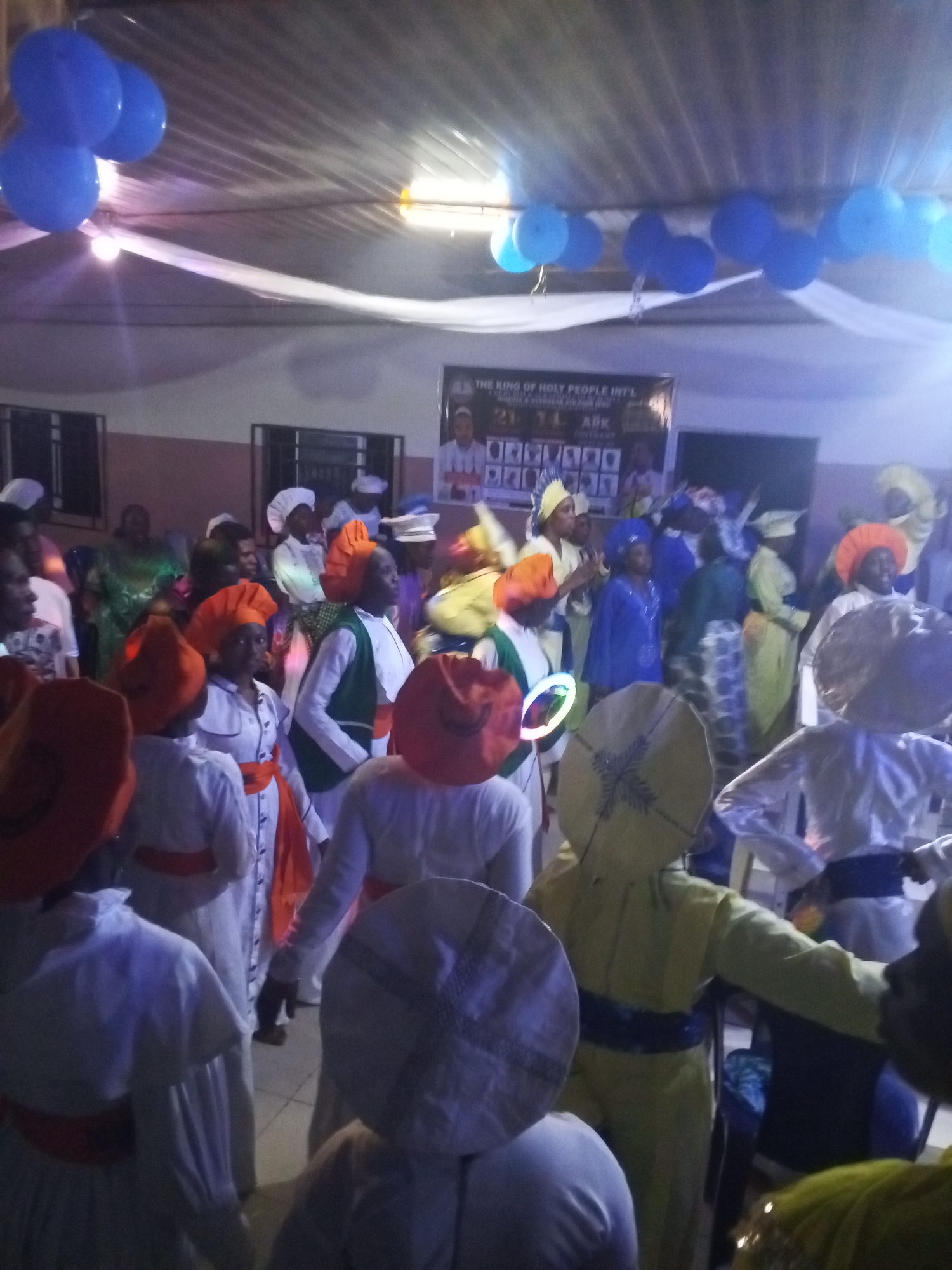 A gathering of people in Lokoja, Nigeria, indoors, seemingly celebrating. The room is decorated with blue balloons and white fabric draping from the ceiling. A banner that reads "THE KING OF HOLY PEOPLE INT'L" is visible. The individuals are dressed in various colored robes and headwear. Some wear orange hats, while others have blue or white coverings. There is a mix of white robes with different sash colors. Some hold circular light-up objects. The scene appears to be festive.