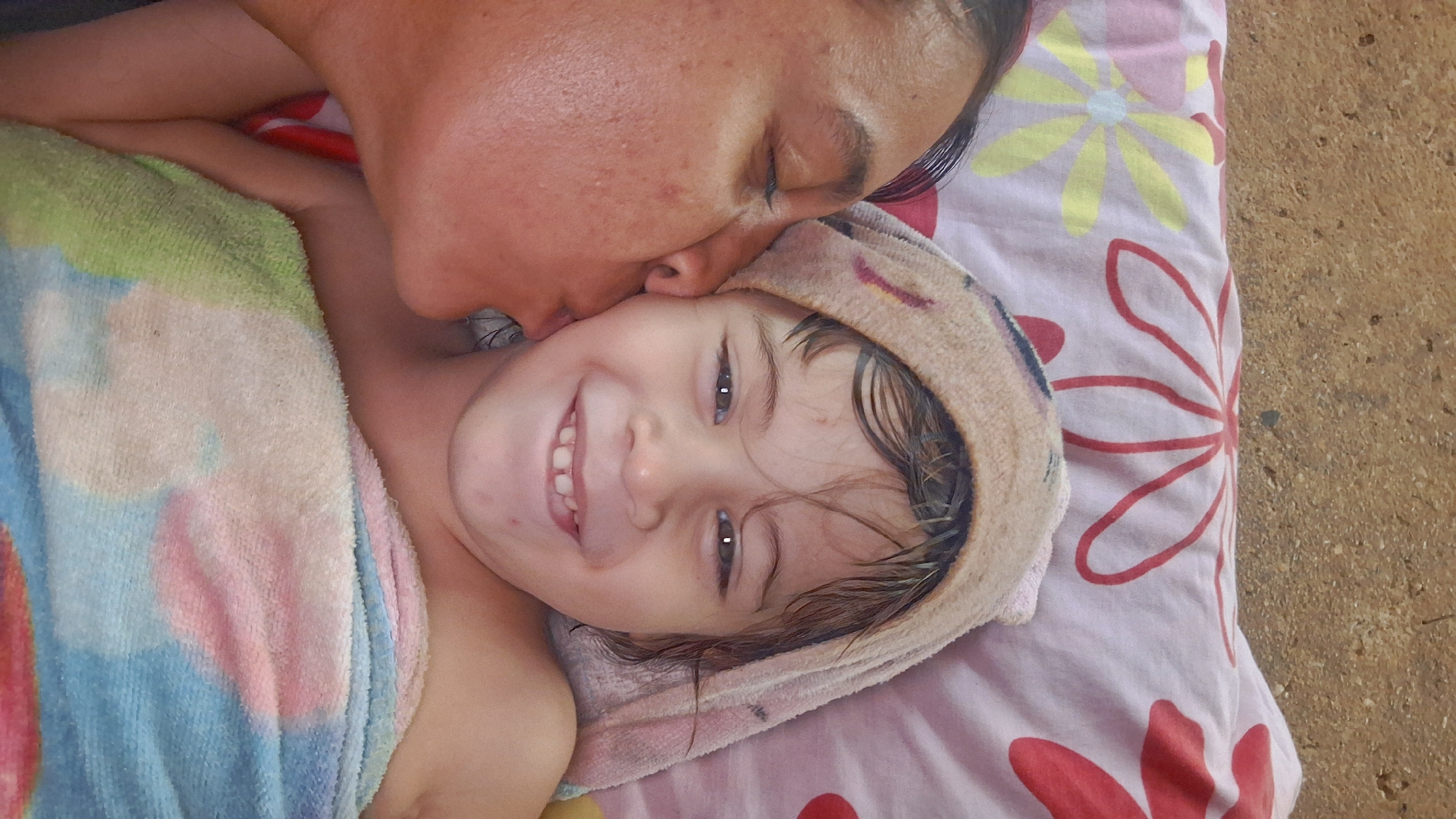 A vertical photograph captures an intimate scene in the city of Vallecito, Venezuela, featuring an adult and a young child. The adult, positioned on the left side of the frame with a medium-brown skin tone exhibiting visible pores and minor blemishes, has their eyes closed and is shown in profile, kissing the child's left cheek with pursed lips. Dark hair is visible near the adult's temple.
The child, centrally positioned and facing forward with a broad smile, has light-brown skin with a few small reddish marks on the forehead and cheek. Dark, wet hair protrudes from beneath a light brown, terry cloth towel wrapped around the child's head. The child's dark eyes are open and directed towards the camera, and their smile reveals upper and lower primary teeth, with a noticeable diastema between the two upper central incisors. The child's body is wrapped in a colorful, tie-dye patterned terry cloth towel, predominantly featuring shades of blue, pink, green, and white.
Both individuals are resting on a light pink fabric surface adorned with stylized floral patterns, including red outlines and yellow petals with light blue centers. Above this patterned fabric, the upper portion of the background consists of a rough, granular, mottled brown and grey surface, consistent with concrete or a similar utilitarian material.