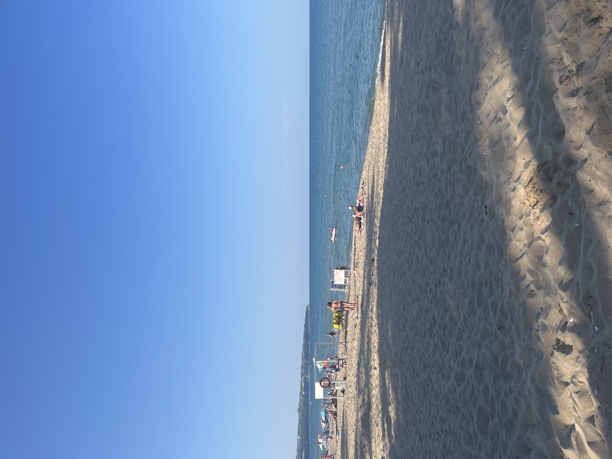 The image depicts a crowded beach scene in Obzor, Bulgaria, under a clear blue sky. The foreground is dominated by the sandy beach, with footprints and shadows cast by unseen trees. In the mid-ground, several people are visible enjoying the sun and sea. Some are sunbathing on towels and loungers, while others are standing or walking along the shoreline. Beach umbrellas provide shade for some individuals.
Further back, closer to the water's edge, a white structure resembling a lifeguard station or information booth is present. A Bulgarian flag is flying from this structure. Adjacent to it are rows of yellow lounge chairs, suggesting a rental service. A few people are interacting around this area.
The sea is a vibrant blue, with gentle waves lapping at the shore. A jet ski is visible in the water to the right, indicating recreational activities. In the distance, a coastline with buildings and greenery can be seen, characteristic of the town of Obzor. The overall atmosphere is one of a typical summer day at a popular seaside resort.
