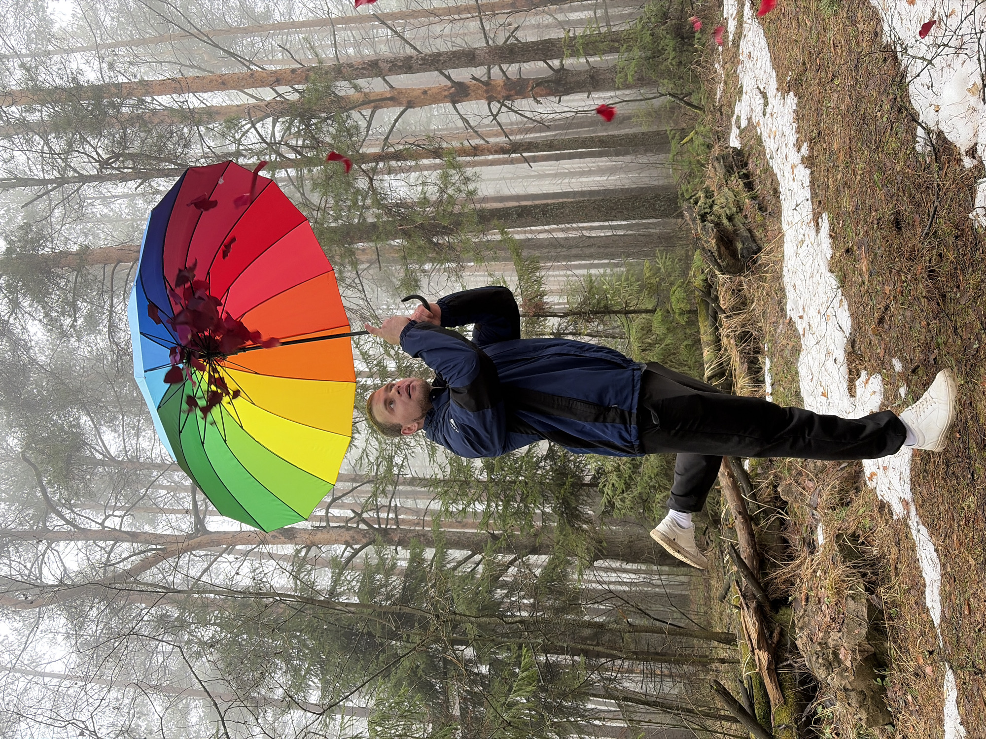 A man stands in a forested area near Прикамский, Russia, holding a rainbow-colored umbrella. The forest is misty, with bare trees and patches of snow on the ground. The man wears a blue jacket, black pants, and white sneakers. He looks up at the umbrella. Red flower petals appear to be falling around the umbrella.