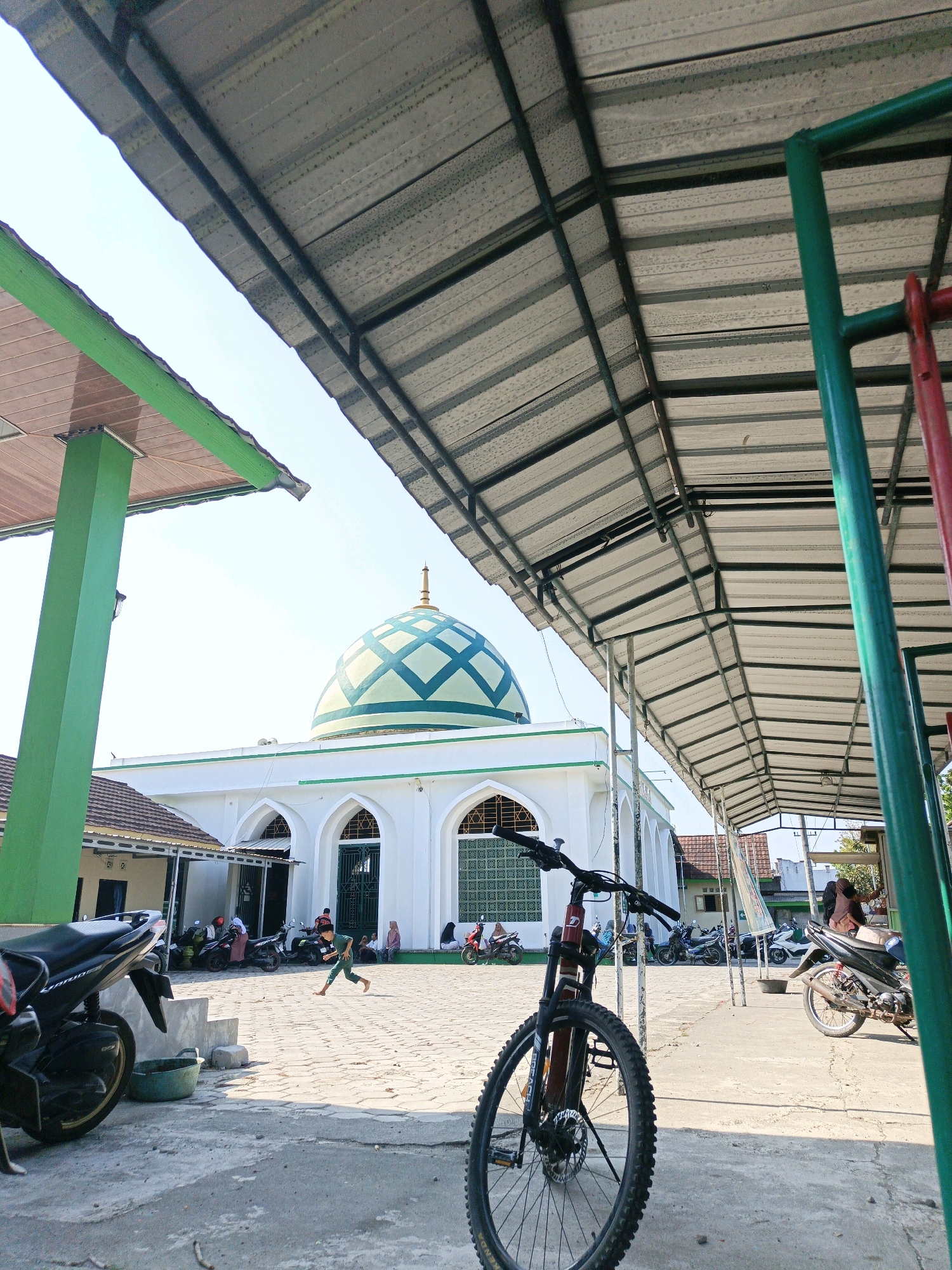 A low-angle, medium shot captures an outdoor scene in front of a mosque in Kalipurwo, Indonesia. The foreground is dominated by the front wheel and handlebars of a mountain bike, angled slightly to the right. Behind the bike, the paved ground is populated by parked motorcycles and several individuals. A young person is in mid-stride, running from left to right across the paved area. Further back, a white mosque with a distinctive teal-and-white patterned dome stands prominently. The mosque has arched windows and doorways. The scene is covered by a metal roof structure supported by green pillars, creating a shaded area with exposed beams and corrugated metal roofing. The sky is clear and bright.