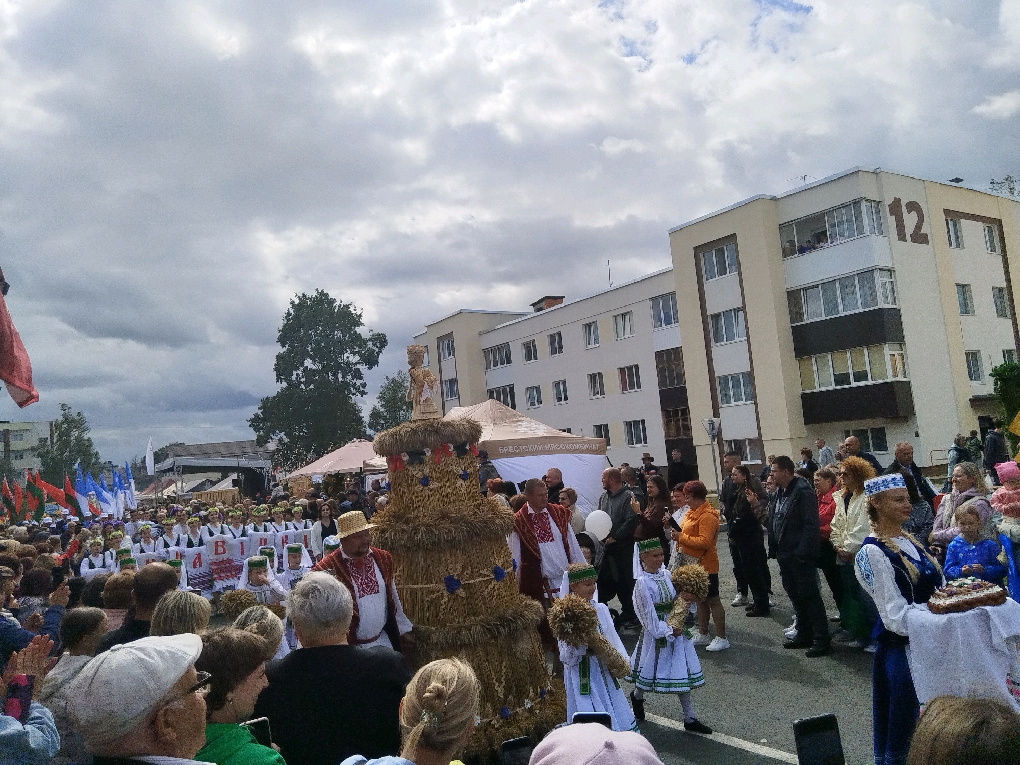 An outdoor daytime shot captures a festive scene with a crowd gathered to watch a procession. In the foreground, a towering, cone-shaped structure made of bundled straw dominates the center, adorned with blue and red decorations. Two young girls in traditional white dresses with green trim walk in front of the straw structure, each holding a small bundle of wheat. A man in a straw hat and a red, embroidered vest also stands near the straw monument. Behind them, a large group of people, some in traditional attire with white headscarves, hold up a banner with Cyrillic lettering. A building with multiple windows and a balcony is visible in the background, with the number "12" prominently displayed on its facade. The sky is overcast with dramatic clouds.