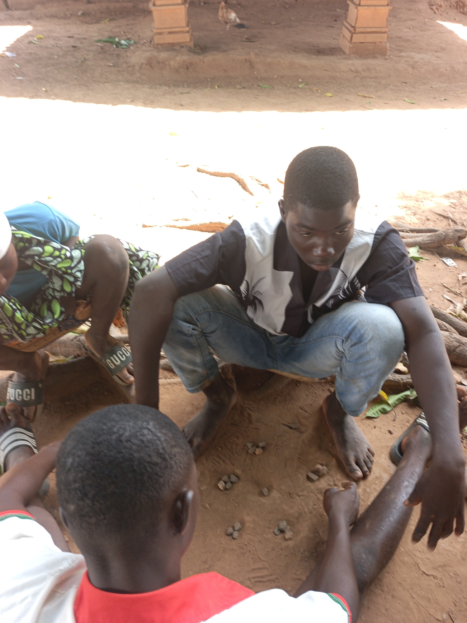 Three people are playing a game in a dirt clearing in what appears to be Akwanga, Nigeria. One person is crouching with bare feet, wearing jeans and a dark shirt with white on the upper chest. Another person wears a green and patterned skirt and Gucci branded sandals. A third person, seen from the back, wears a white shirt with red collar and sleeve details. Small stones are arranged in the dirt in front of them. A chicken stands in the background, partially obscured by shade.