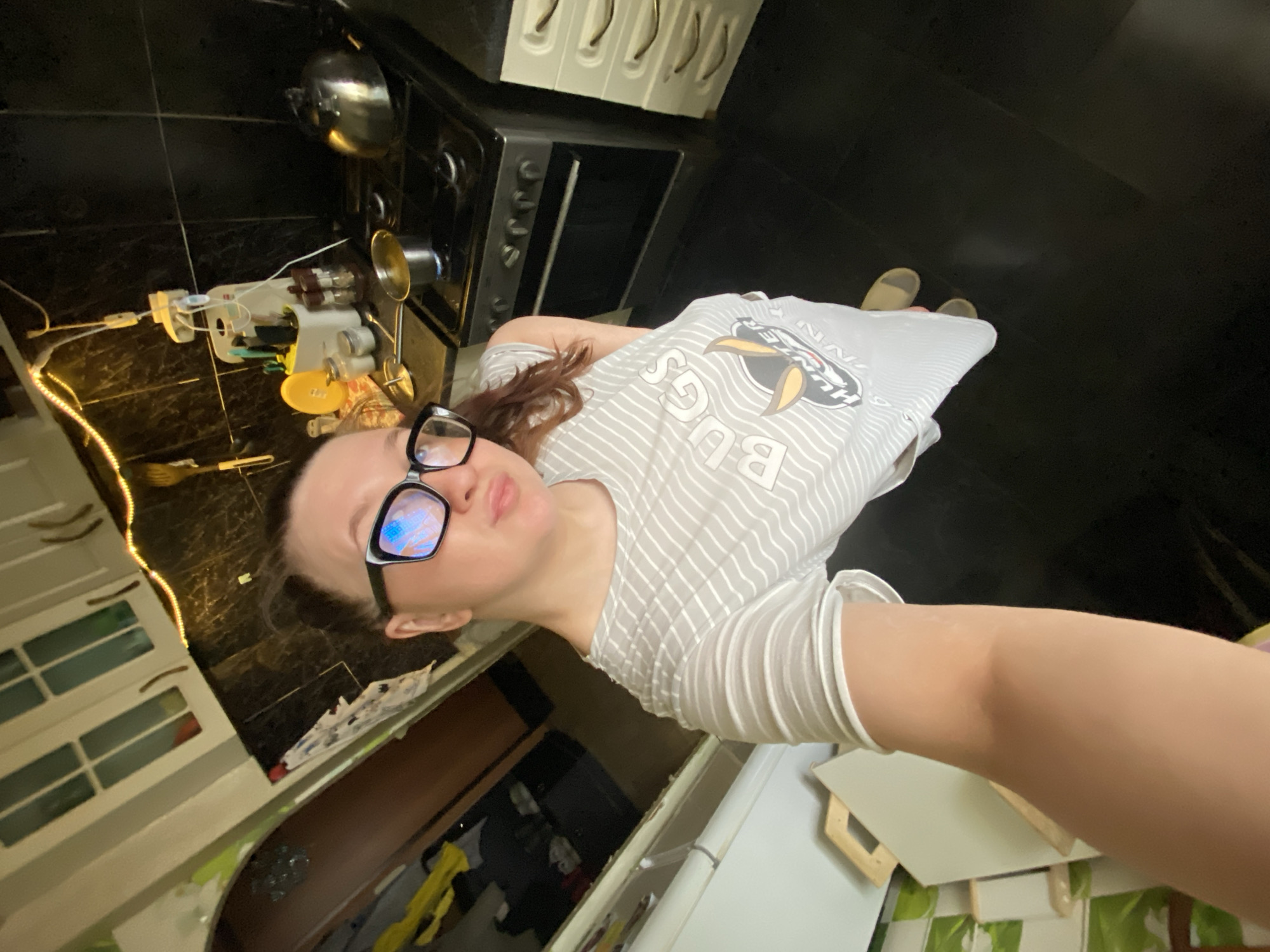 A woman wearing glasses and a gray and white striped shirt with a "BUGS HUNTER" logo is taking a selfie in a kitchen. The kitchen has black tile flooring, white cabinets with silver handles, a black stove, and a refrigerator. A spice rack and hanging pots are visible above the stove. String lights are affixed to the cabinetry. The specific location and city of Chaikovsky, Russia, cannot be confirmed from this image alone.