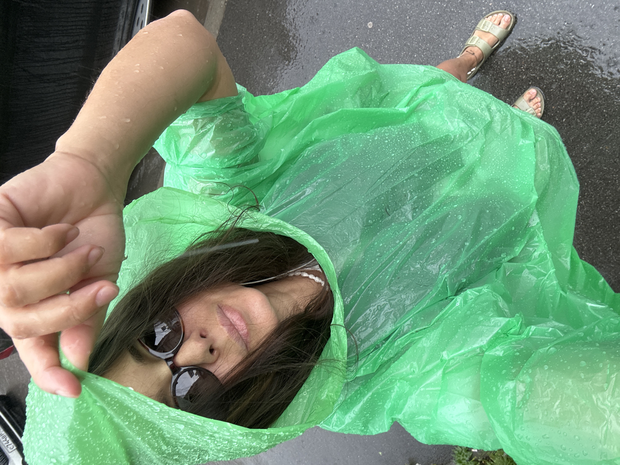 A person is wearing a bright green, crinkled, disposable poncho that is speckled with raindrops. The poncho covers most of their body, with only their face, arm, and feet visible. The person is looking up and to their right, with a slight smile. They are wearing dark sunglasses and white earbuds. Their arm is raised and their hand is near the hood of the poncho, pulling it slightly away from their face. The person is wearing light-colored sandals on their feet, and there are water droplets on their skin and the poncho.
The scene appears to be outdoors, with a dark, wet surface resembling pavement or asphalt visible in the background and on the ground. The presence of raindrops on the poncho and the wet ground strongly indicate that it is raining. The lighting suggests it is daytime, but the overcast sky due to the rain likely makes it feel dim. The image is taken from a high-angle perspective, looking down on the person. There are no visible landmarks that would pinpoint the location within Kazan, Russia, but the overall scene is consistent with an urban or semi-urban environment during a rainy day. The person's expression is relaxed and perhaps even slightly amused, despite the rain. No text is visible in the image.