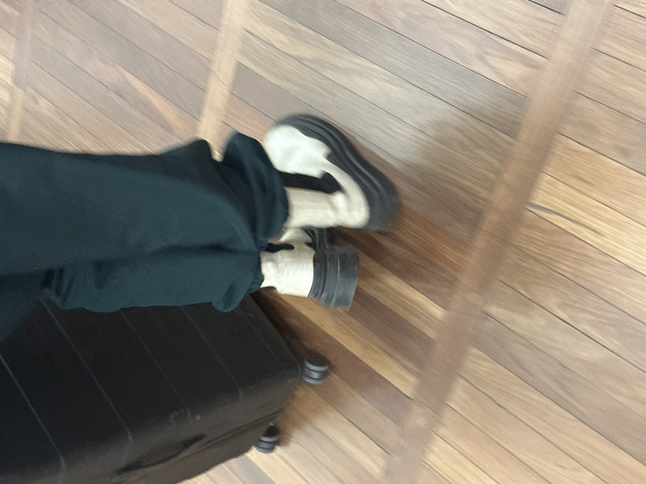 The lower half of an individual is depicted in an indoor setting. The person wears dark green trousers and light-colored, possibly beige or off-white, chunky-soled boots with thick black platforms. A black hard-shell rolling suitcase with four black wheels is positioned to the left of the individual's left leg. The individual appears to be in motion, suggested by slight blur, seemingly walking forward while accompanying the suitcase. The floor is composed of polished wooden planks, exhibiting variations in shade from light to medium brown. The specific location within London, United Kingdom, cannot be determined from the provided image.