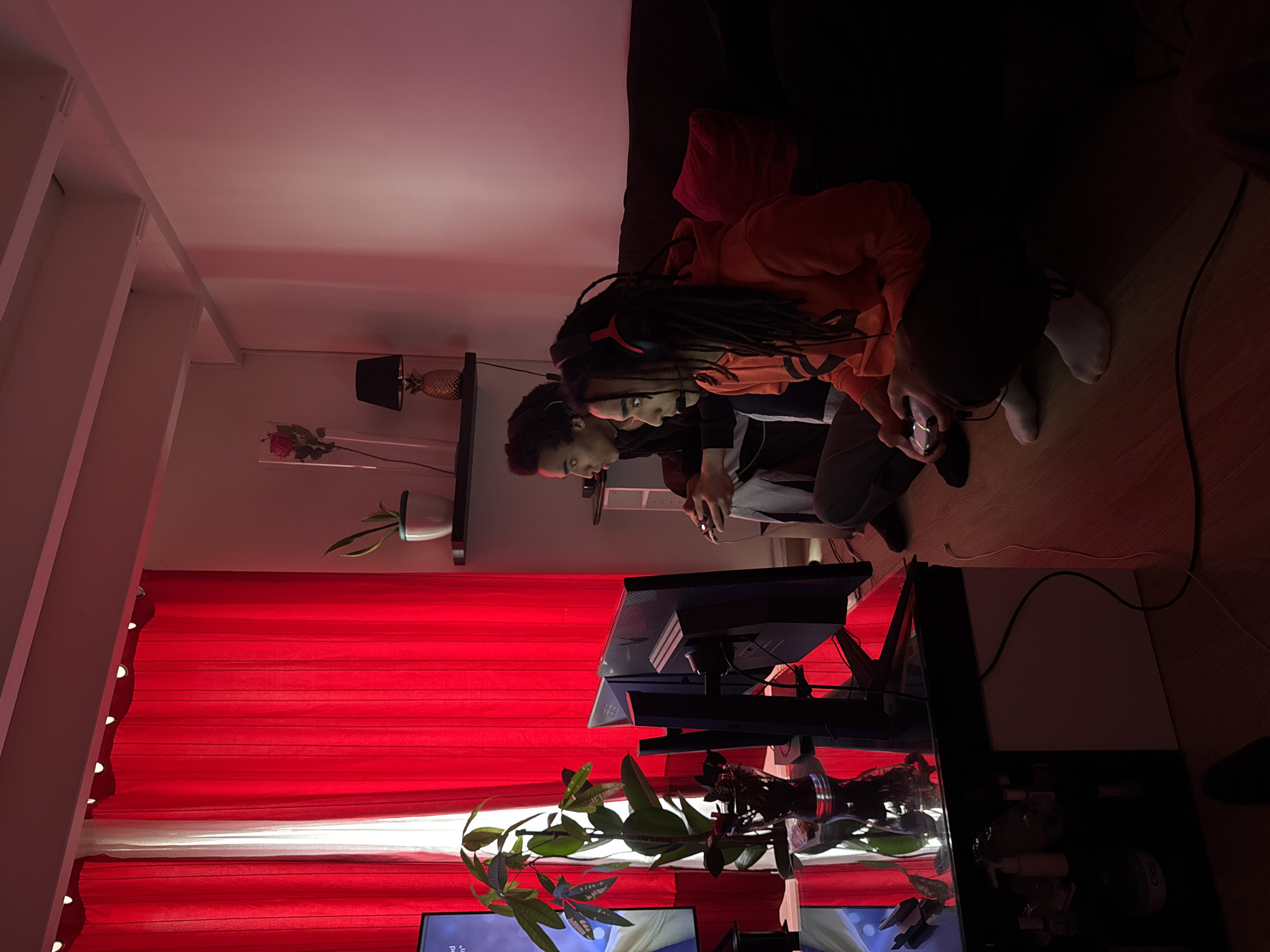 Two individuals are seated side-by-side on a dark-colored couch, engaged in video games. The person in the foreground is wearing an orange hoodie and has dreadlocks, adorned with a gaming headset. The other person, partially obscured, is wearing a dark-colored top. Both are holding game controllers, focused on the screens in front of them.
In front of them, there is a desk with multiple monitors and gaming equipment. A houseplant with broad green leaves sits in front of the monitors. Red and white vertically-striped curtains hang in the background. On a wall-mounted shelf, there is a black lamp, a decorative pineapple, and potted plants.
The location cannot be determined to be Paris, France.