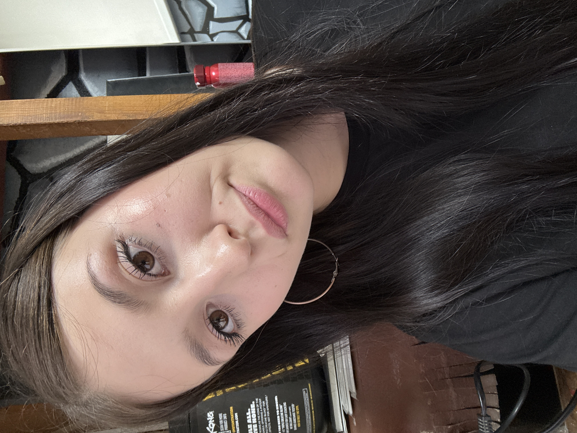The image is a close-up, selfie-style shot of a young woman with long, dark hair and light brown eyes. She is wearing a black t-shirt and a large hoop earring in her visible ear. Her makeup includes mascara and pink lipstick. She is looking towards the camera with a slight smile. In the background, there is a black container with yellow labeling on the left, a wooden structure, a wall with a geometric pattern, and a small red object. The lighting suggests an indoor environment. There is no discernible information in the image that would indicate its location in Chaikovsky, Russia, or any other specific city.