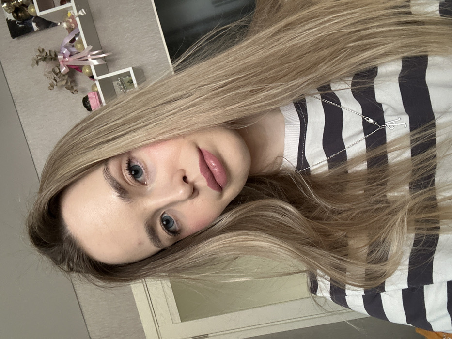 A young fair-skinned female with long, straight blonde hair and blue eyes is depicted in a close-up, chest-up shot. Her gaze is directed towards the camera. She wears a long-sleeved shirt with alternating white and dark grey horizontal stripes. A silver-colored chain necklace with a cursive letter 'A' pendant rests on her chest. Her lips are coated with a pinkish gloss, and a light blush is visible on her cheeks. The background, indicative of an indoor setting in Chaikovsky, Russia, features a light-colored, possibly textured wallpaper. To the subject's left, a portion of a light-colored door frame is visible. To her right, a white multi-compartment wall shelf is affixed, displaying various decorative items including a dried floral arrangement with pink ribbon, small spherical ornaments, a small pink object, and a rectangular bottle. Below the shelf, a dark, rectangular screen, potentially a television, is partially visible.