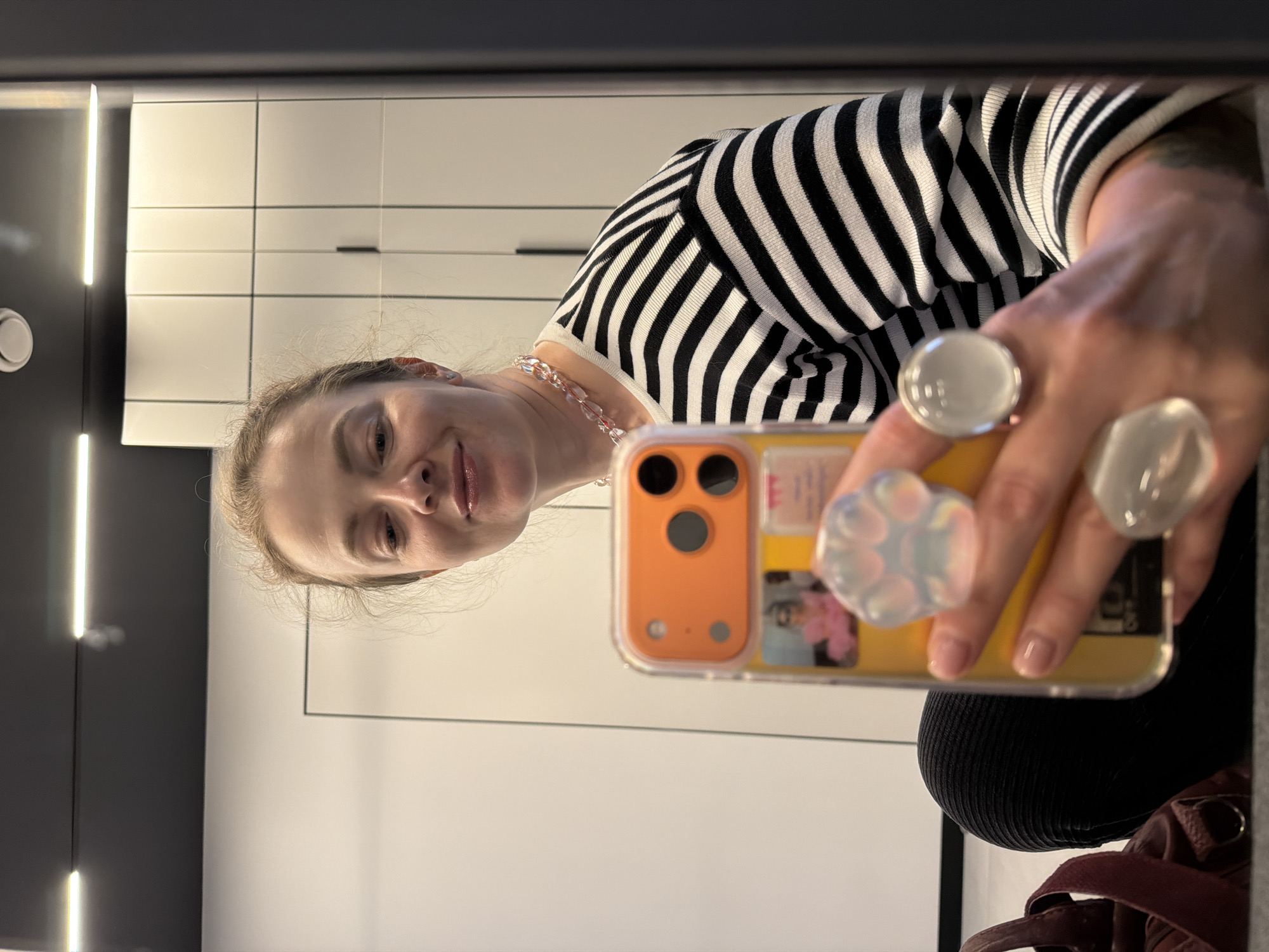 A woman is taking a selfie in a mirror. She is wearing a black and white striped shirt and a pink beaded necklace. She is holding an orange smartphone. She is wearing two large rings with white stones. In the background, there are white cabinets with black handles. It is not possible to determine the location in the city of Moscow, Russia from the given information.