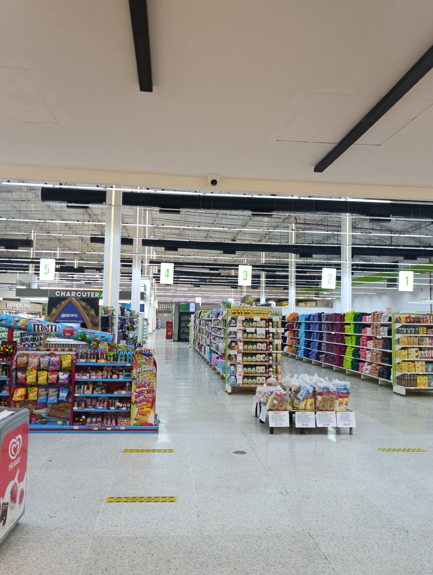 The image shows the interior of a supermarket, likely located in Porlamar, Venezuela. A wide aisle stretches into the distance, marked by numbered signs hanging from the ceiling (5, 4, 3, 2, 1). Shelves are stocked with various items. The left side of the aisle is filled with displays of candy, chips, and snacks. Further down on the left is a sign that reads "CHARCUTERIA" and "REFRIGERADOS". On the right side, there are shelves of goods including towels. In the middle aisle, stands an island of sacks of rice or grain with price labels. The floor has markings indicating social distancing (1 metro minimo). The ceiling has a network of lights and structural elements. A security camera is visible. There are no people clearly visible in the image.