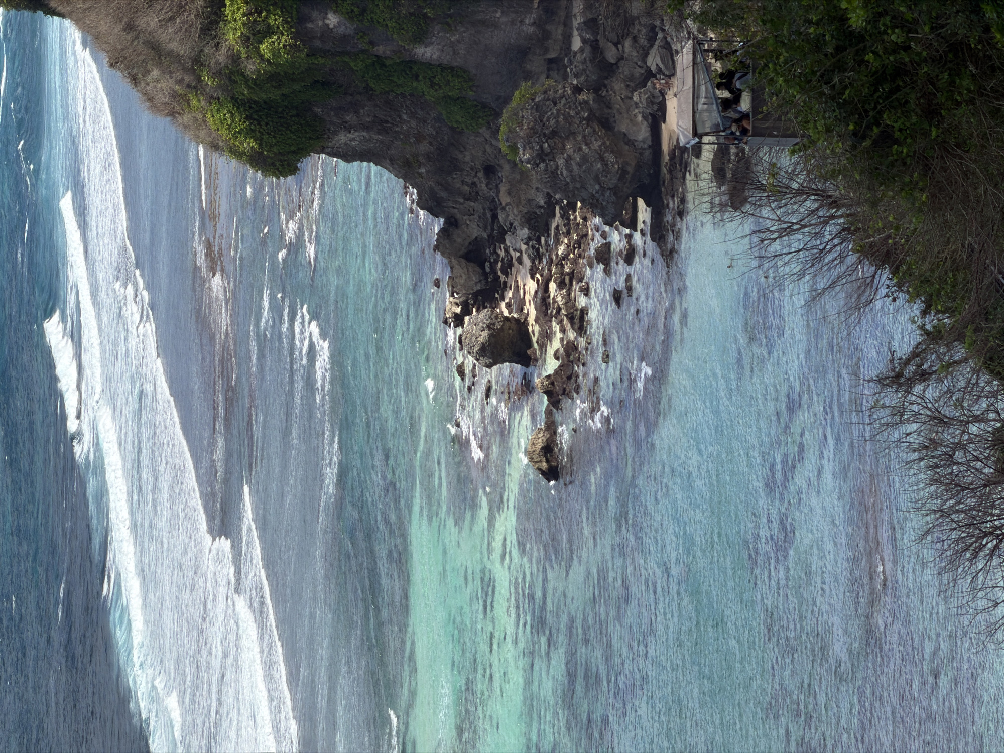 This image shows a coastal landscape with a cliff overlooking the ocean. The water is a clear turquoise near the shore, gradually deepening to a darker blue further out. Waves are breaking on the shore, creating white foam. The cliff is covered in lush green vegetation in some areas and appears dry and rocky in others. There are several large boulders and rocks along the coastline at the base of the cliff.
In the lower right portion of the image, there is a covered structure that appears to be a viewing platform or shelter. Inside the shelter, several people are visible, some appearing to be seated. Two individuals are standing on a sandy area near the shelter, closer to the water's edge. The vegetation in the foreground includes dense green bushes and the bare branches of trees. The scene is consistent with the coastal areas of Kuta Selatan, Indonesia, known for its dramatic cliffs and beautiful ocean views.