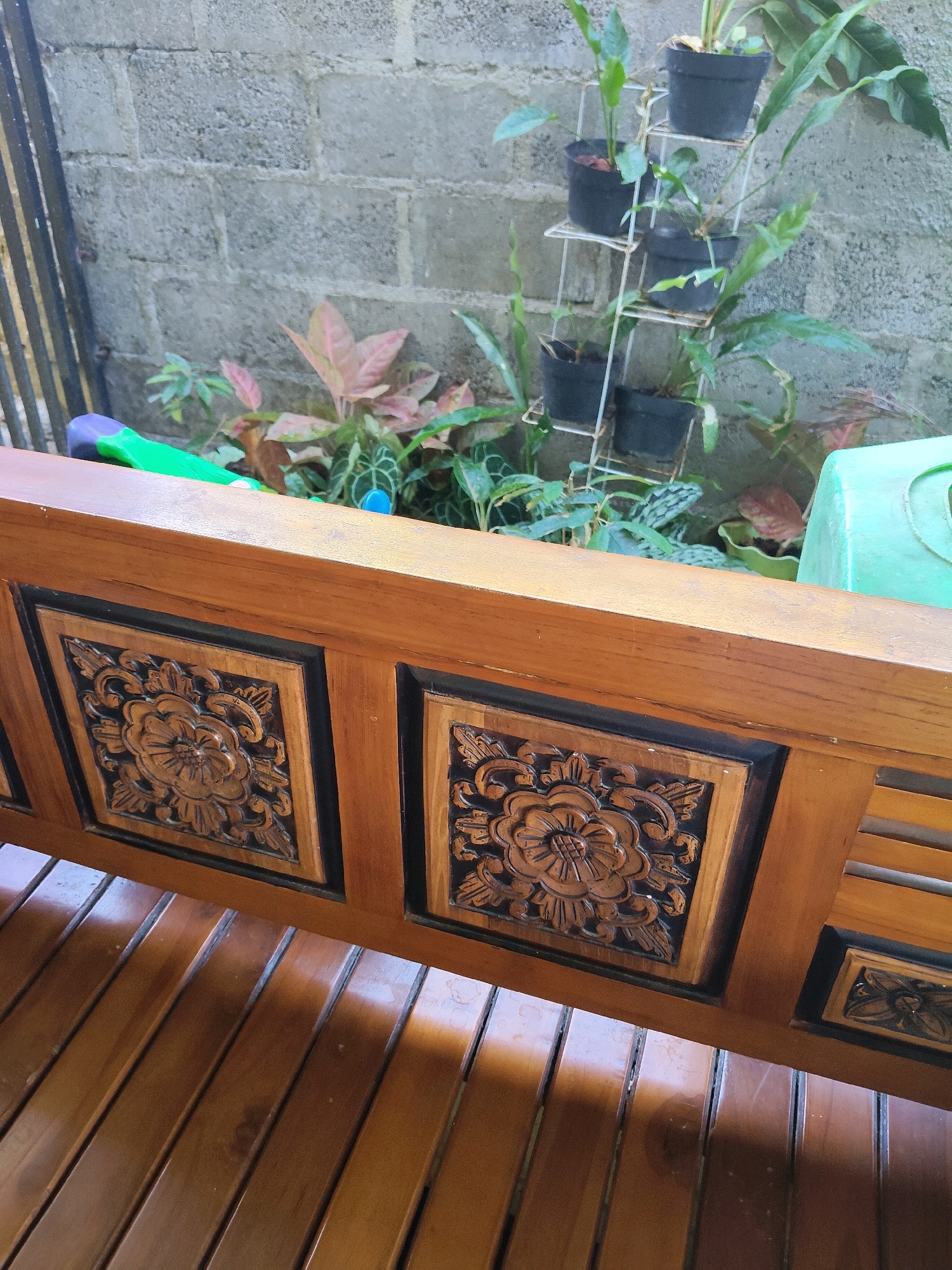 The image depicts a close-up view of a wooden bench with intricate floral carvings on its backrest, set against a backdrop of a brick wall and several potted plants. The bench is made of polished wood, with slats forming the seat. The backrest features square panels with carved floral motifs, outlined in black. Behind the bench, a wall constructed from grey cinder blocks is visible. To the left of the wall, a portion of a dark metal fence is present. A multi-tiered metal plant stand holds several black pots containing various green plants. Among the plants, a green and purple object, possibly a garden tool, lies on the bench. To the far right, a portion of a light green plastic object, likely a container, is visible. The scene appears to be an outdoor area, possibly a patio or garden, and is likely located in Kupang, Indonesia, given the characteristic brick wall construction and tropical plant types common in the region. There are no people visible in the image.