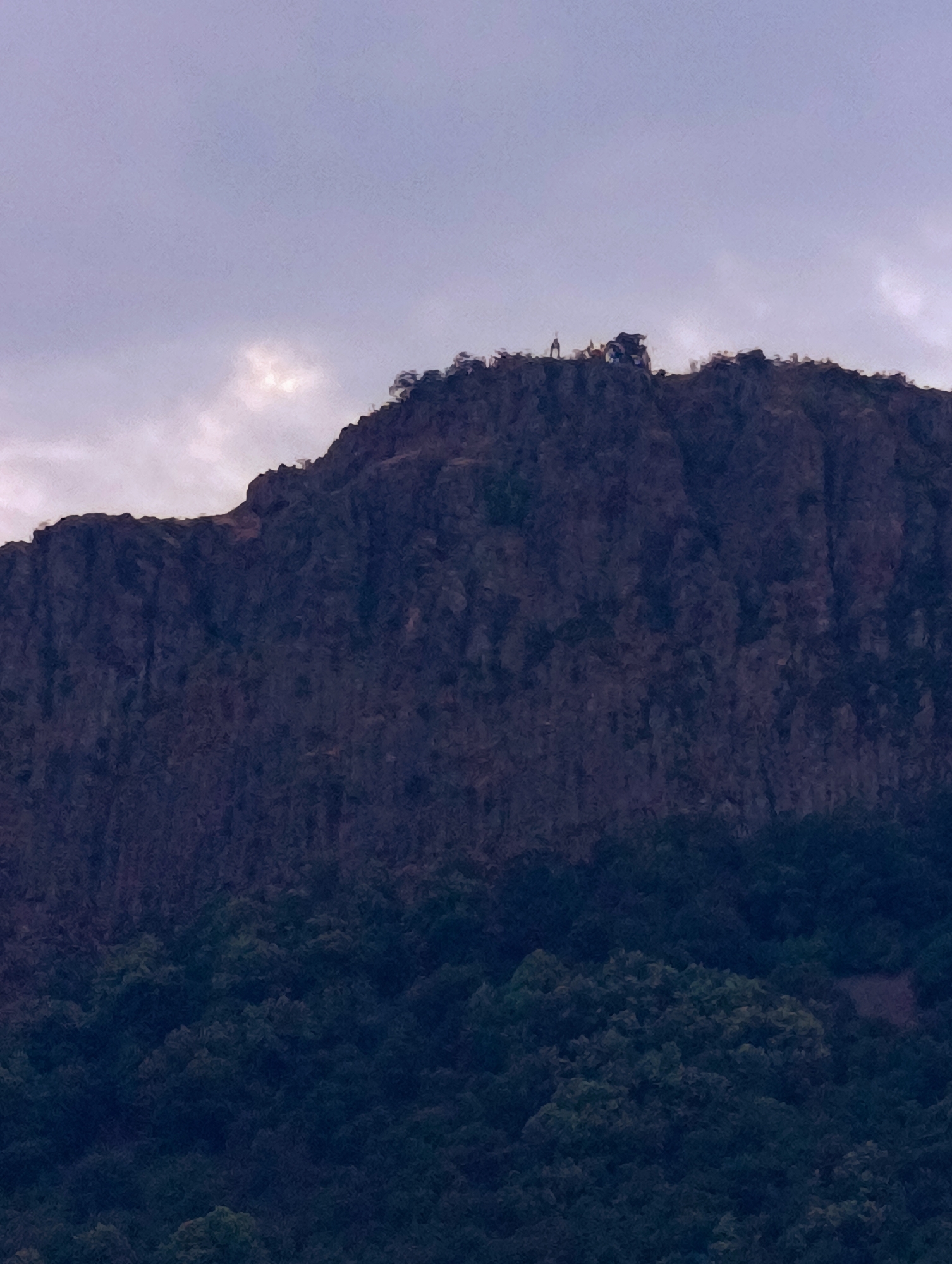 The image shows a rocky mountain peak with some trees at its base. Two figures stand on the very top of the mountain, silhouetted against a cloudy sky. The sky is light purple and grey with some patches of brightness. The mountain is dark and textured, with the lower part covered in a dense green forest. The image appears to be a photograph taken from a distance, capturing a dramatic natural landscape. The location is Shiroko Pole, Bulgaria.