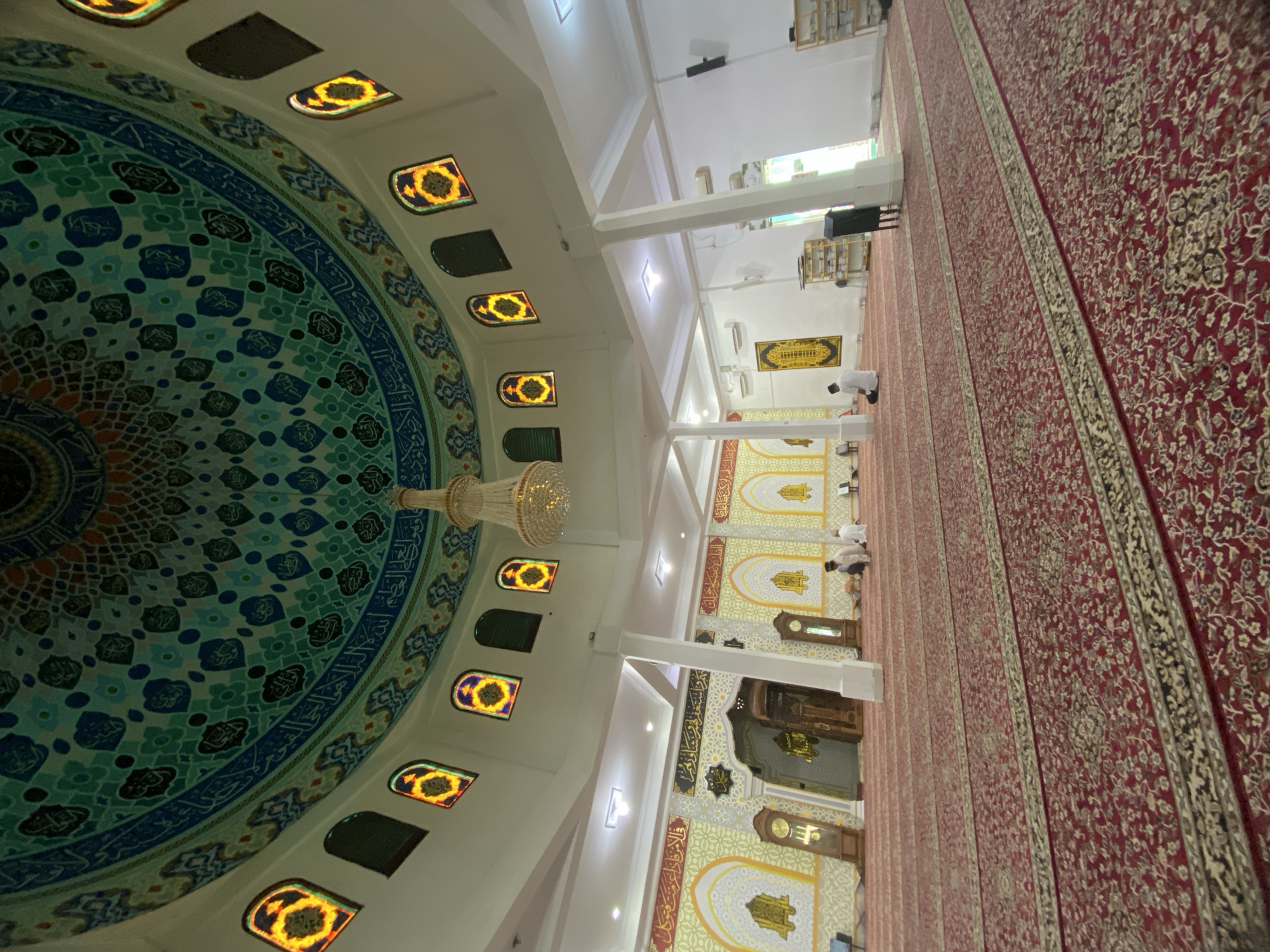 The image depicts the interior of a mosque, likely the Masjid Agung Islamic Center in Lhokseumawe, Indonesia, given the architectural style and decorative elements. The focal point is the ornate, domed ceiling, adorned with intricate patterns in shades of green, blue, and black, interspersed with Arabic calligraphy. Surrounding the dome are arched stained-glass windows featuring a vibrant yellow and orange floral motif. A large, multi-tiered crystal chandelier hangs from the center of the dome.
The main prayer hall is spacious, covered with a large, intricately patterned red carpet with prominent borders. Several individuals are present, dressed in white attire, engaged in prayer or contemplation. Some are kneeling on the carpet, facing the mihrab, a niche in the wall indicating the direction of Mecca. The walls of the prayer hall are decorated with geometric patterns and calligraphy, framed by arches. Tall, wooden clock towers are visible on either side of the entrance to the mihrab area, which is ornately carved. The environment appears well-lit, with ceiling lights and natural light filtering through the windows. The overall impression is one of serenity and devotion.