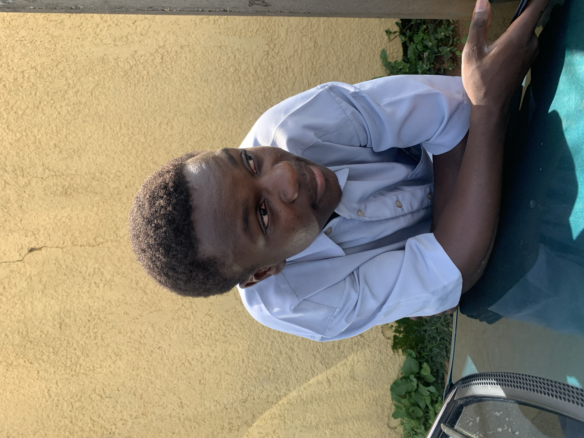 A young Black man is leaning on the hood of a dark green car, with his arms crossed. He is wearing a light blue collared shirt. He is looking to his right, with his head slightly tilted. The background consists of a textured yellow wall, with some green foliage at the bottom left. A portion of a concrete pole is visible on the right side of the image. The scene appears to be outdoors, possibly in a residential area in Jos, Nigeria, given the architectural style of the wall and the presence of a car.