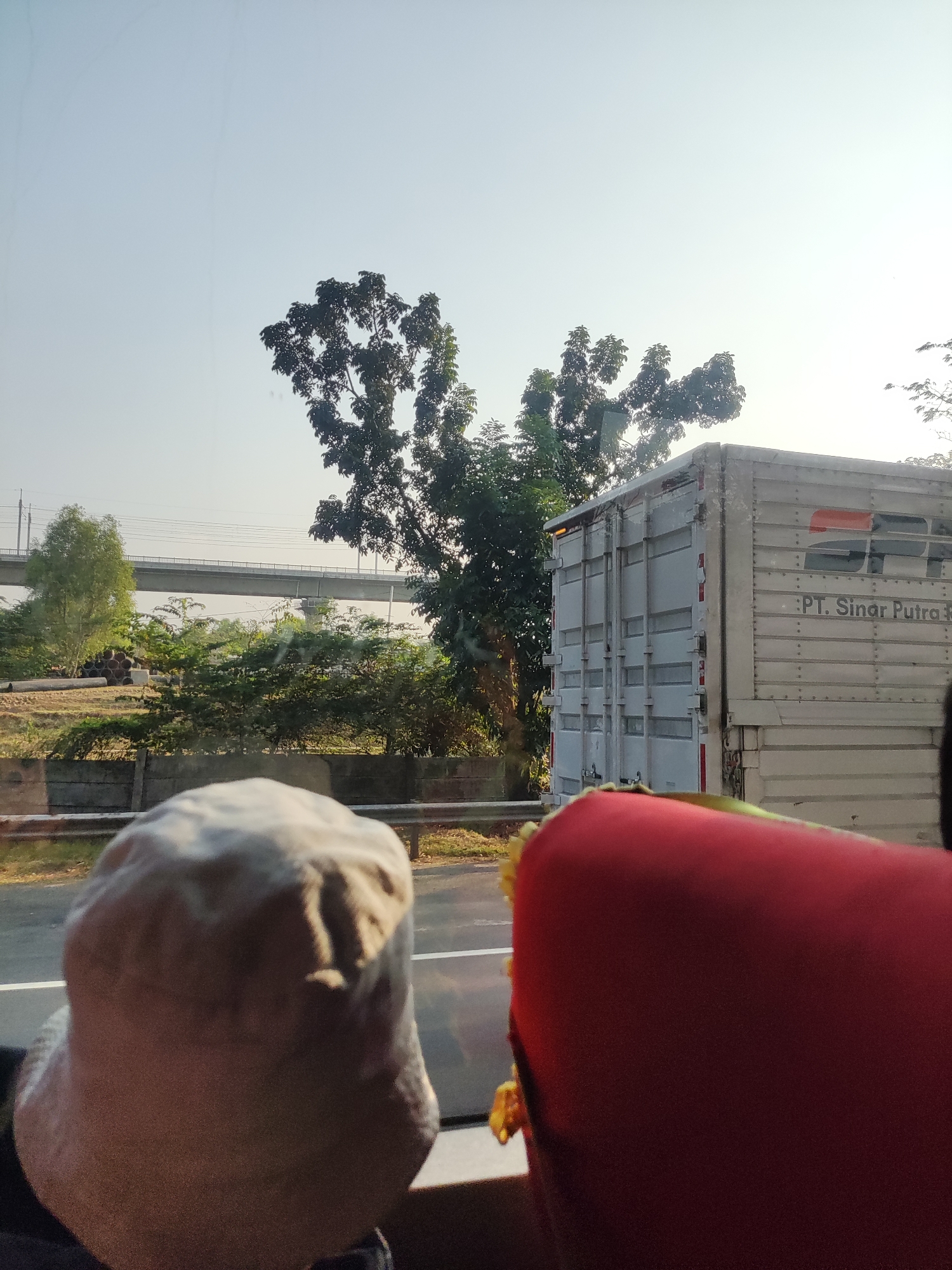 The image captures a view from inside a vehicle, looking outwards towards a road and its surroundings. In the foreground, the top of a person's head, covered by a light-colored hat, is visible on the left. To the right, the back of a red seat or cushion is prominent.
The midground features a light-colored box truck with the logo of "PT. Sinar Putra." The truck is parked or moving alongside the road. Adjacent to the truck and the road, there is a concrete barrier and vegetation.
In the background, a large elevated bridge structure, likely part of a highway or railway, spans across the scene. Trees and other foliage are visible, suggesting a semi-urban or rural environment. The sky is clear and bright, indicating daytime. Based on visual cues such as the vegetation and architecture, the location is identified as Hegarmukti, Indonesia. The overall impression is a snapshot taken during a journey, possibly on a highway.