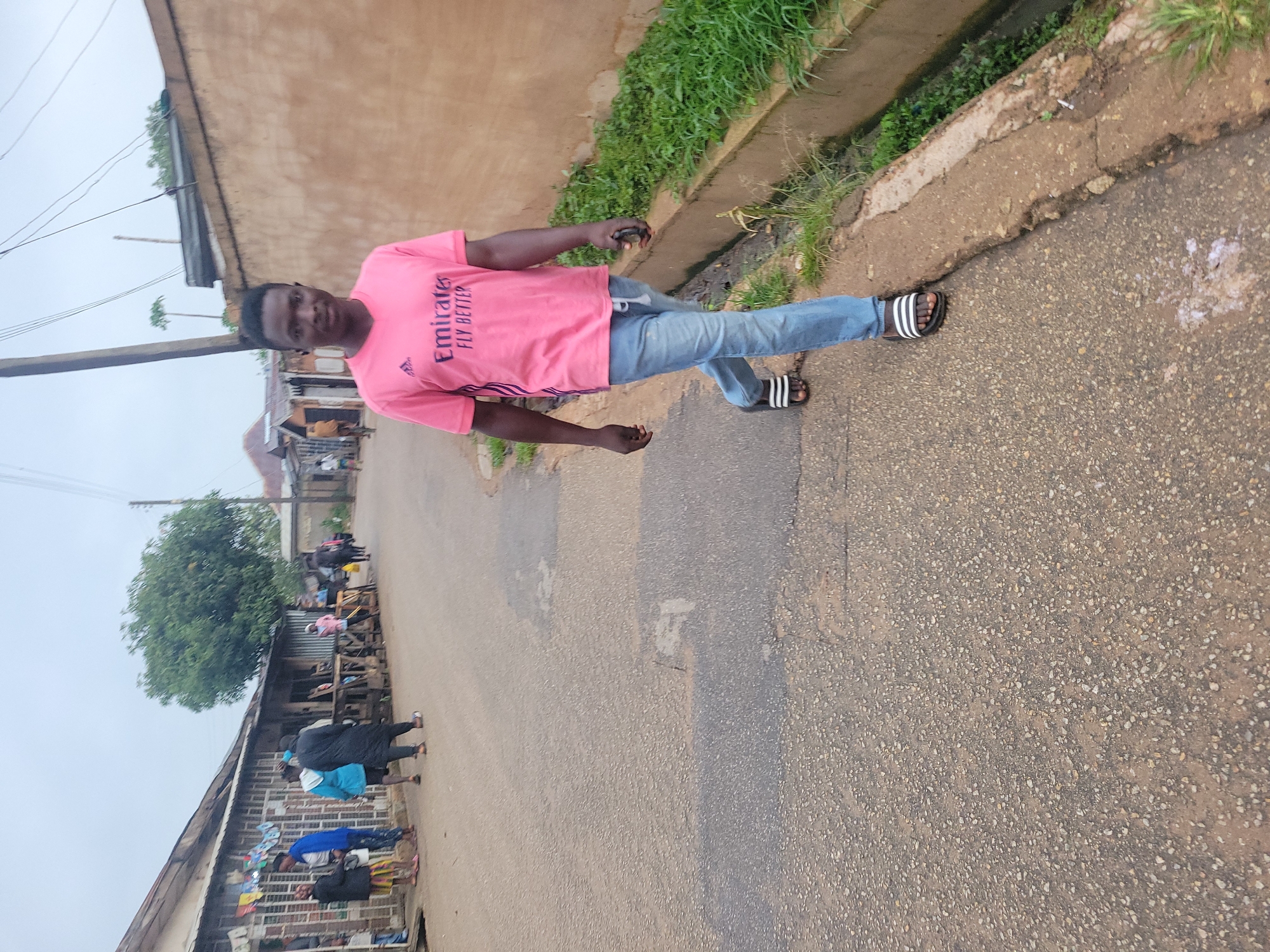 A young Black man wearing a bright pink t-shirt with "Emirates FLY BETTER" printed on it and light blue jeans walks down a paved street. He is wearing black and white striped slides. His expression is neutral as he looks towards the camera. He is walking past a drainage ditch filled with green plants and a textured, tan wall. In the background, other people are visible near buildings that appear to be shops or homes, suggesting an urban environment in Jos, Nigeria. The sky is overcast.