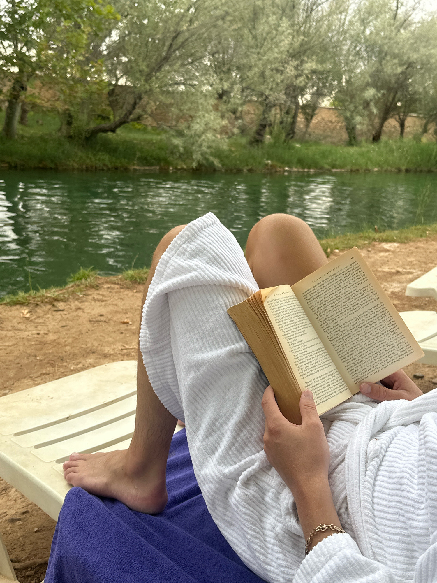 A person, partially visible from the waist down, sits on a lounge chair next to a body of water. They are wearing a white robe and reading a book. Their bare feet rest on the chair and a purple towel. The environment appears to be a natural setting with trees and vegetation lining the water's edge. The person's hand is visible holding the book, and they are wearing a bracelet on their wrist. The lighting suggests it is daytime. This scene could be set in the spa town of Alhama de Aragón, Spain, known for its thermal waters and natural beauty.