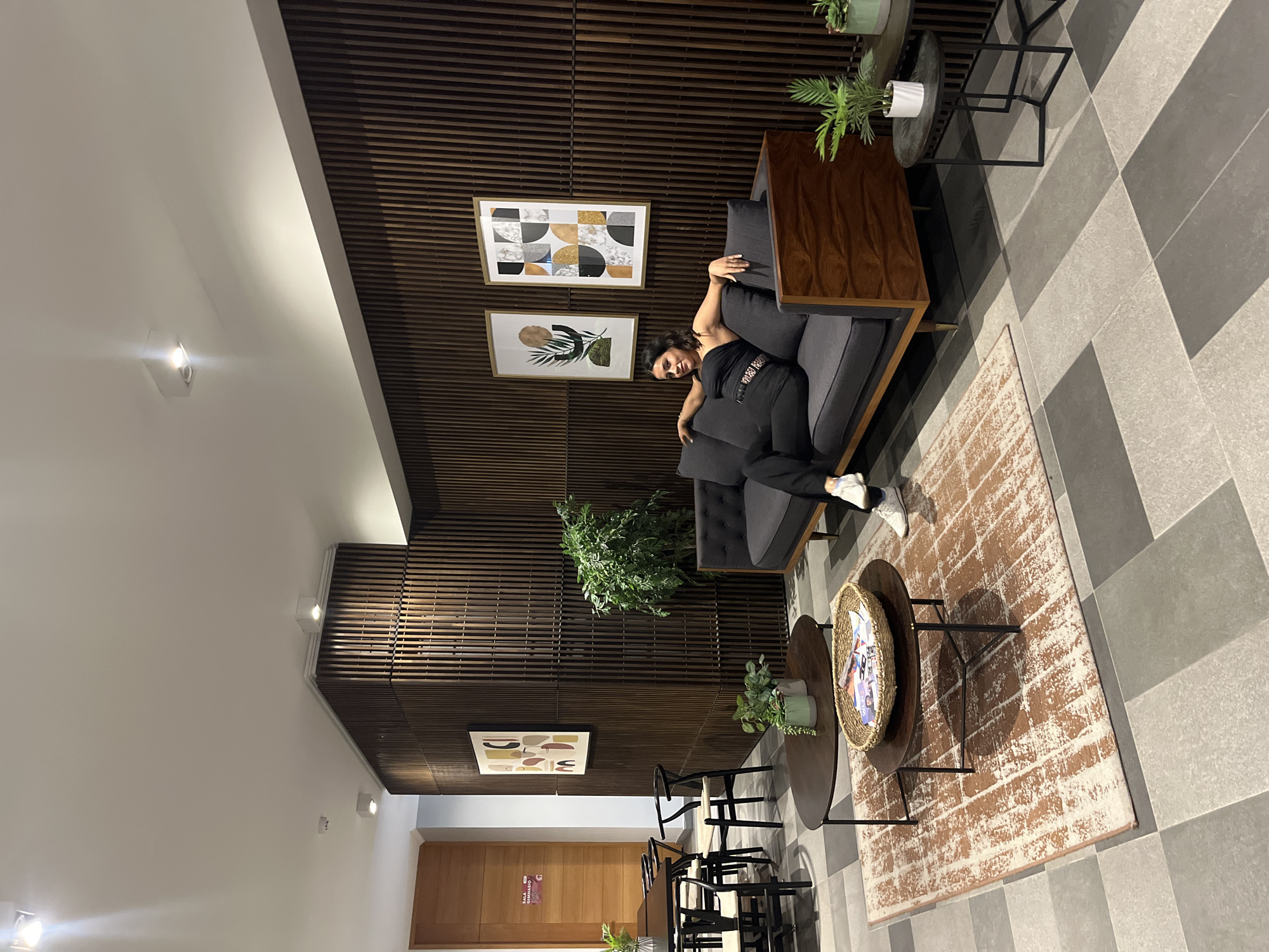 The image depicts the interior of a modern space, likely a lobby or waiting area. A person is reclined on a gray sofa, posing for the camera. The walls are paneled with dark wood slats, and artwork hangs on the wall behind the sofa. A patterned rug covers the floor near a low, round coffee table. Several chairs are placed around a second round table. Potted plants are scattered throughout the space, adding greenery. The overall aesthetic is contemporary and stylish. The location appears to be in an Unknown City, Unknown.
