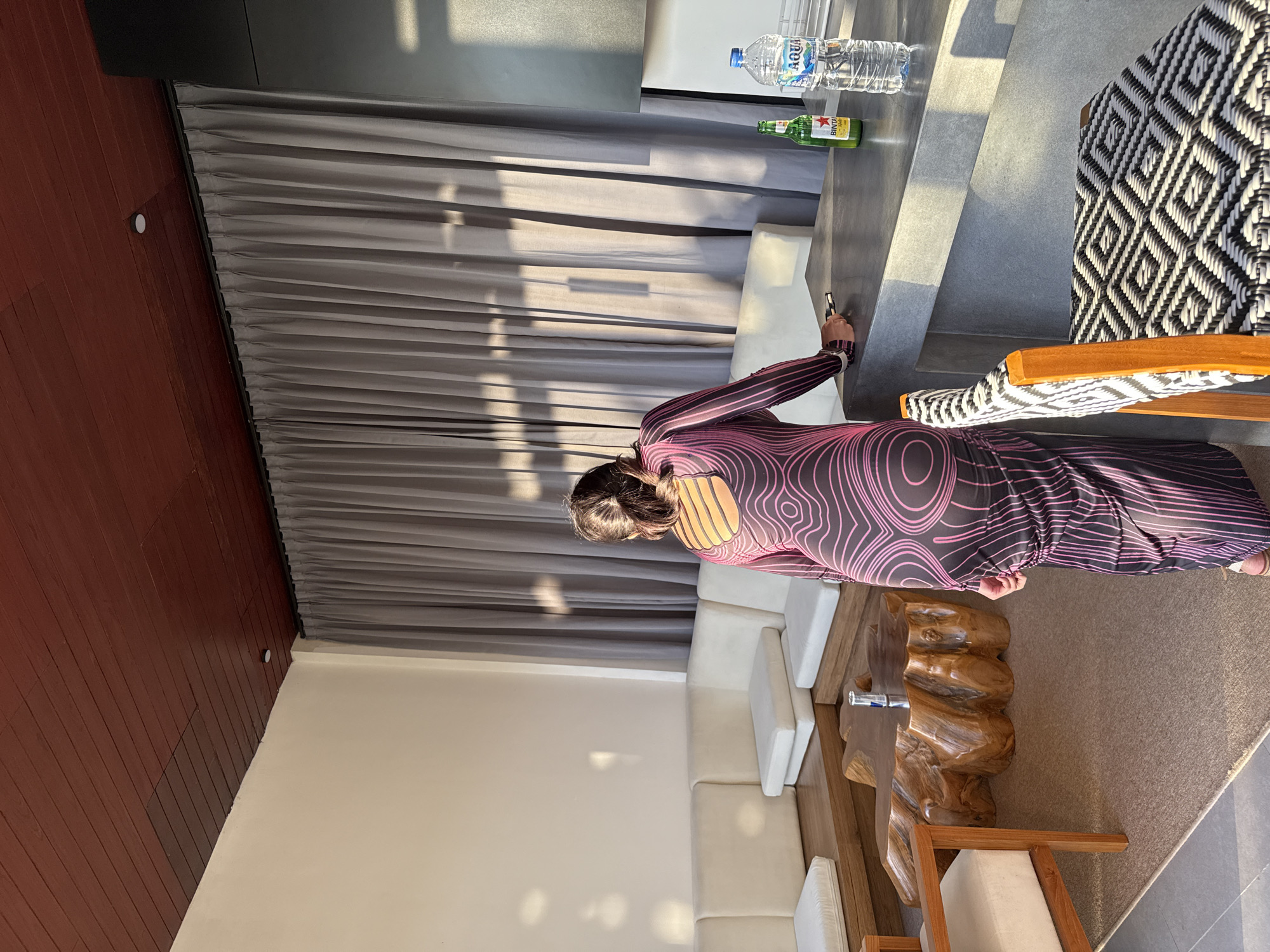 A woman wearing a long, form-fitting, dark-colored dress with a pink wavy line pattern stands facing away from the camera, her back to the viewer. She is leaning against a grey countertop, holding a dark object in her right hand. To her right on the countertop are a green bottle of Bintang beer and a clear bottle of Aqua water. Behind her, a floor-to-ceiling grey curtain is drawn, and sunlight casts shadows on it.
In the foreground, a patterned bar stool with a wooden frame and a black and white geometric woven seat is visible. The environment appears to be an indoor lounge area with a large, light-colored sectional sofa and a unique, irregularly shaped wooden coffee table. The ceiling is made of dark reddish-brown wood planks. The overall setting suggests a modern, possibly resort or residential, space in Kediri, Indonesia.