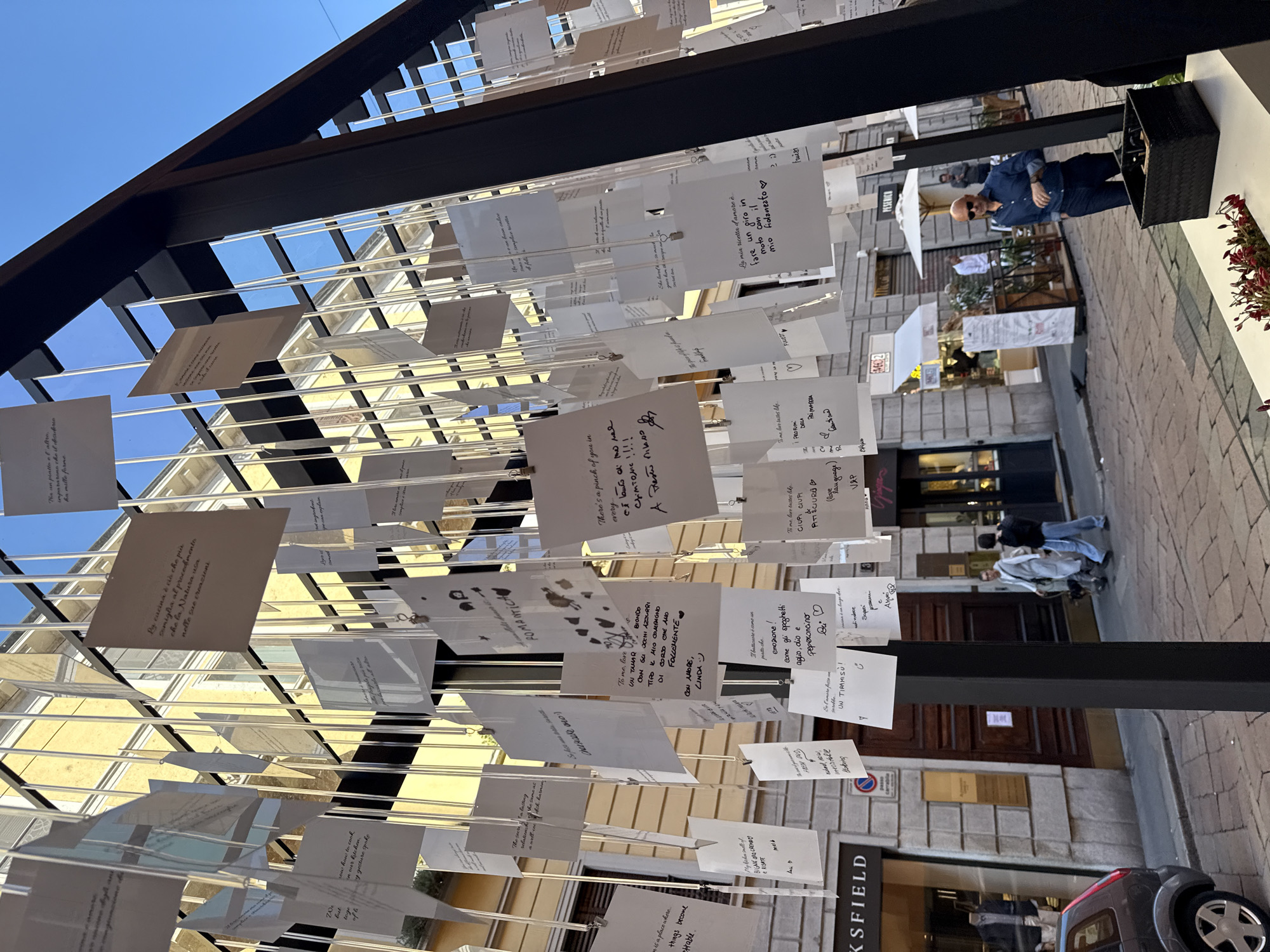 The image shows an outdoor art installation in Milan, Italy. A black metal framework supports numerous pieces of paper suspended from clear plastic lines. Handwritten notes and drawings are visible on the papers. The structure is set on a paved street in a commercial area, with buildings on either side. A man in a blue shirt stands in the background. A grey car is parked in the lower right corner.