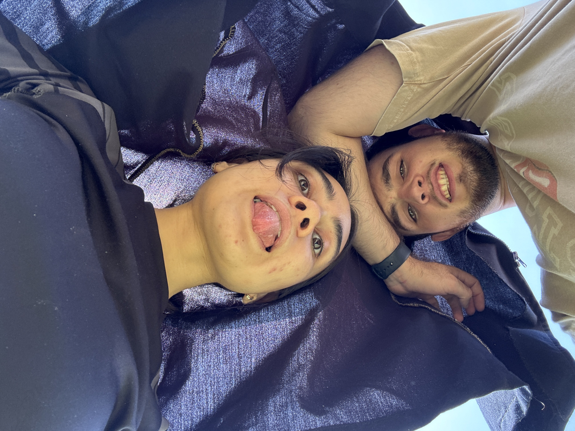 Two people lie on a shiny, dark purple fabric. A person on the left wears a black top and sticks their tongue out. A person on the right wears a light brown t-shirt and smiles. The background is a clear sky. The location is unknown.