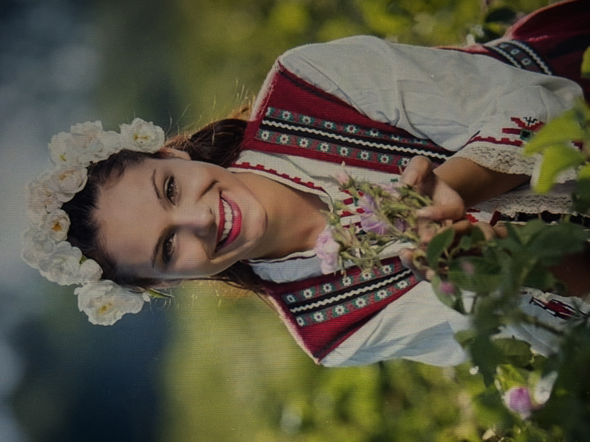 A young woman with a flower crown smiles at the camera, holding a small bouquet of pink roses in her hands. She wears a traditional white blouse with an embroidered red vest over it. The background is a field of green plants, suggesting an outdoor rural setting. The lighting is soft and natural, with the sun highlighting the woman's face.