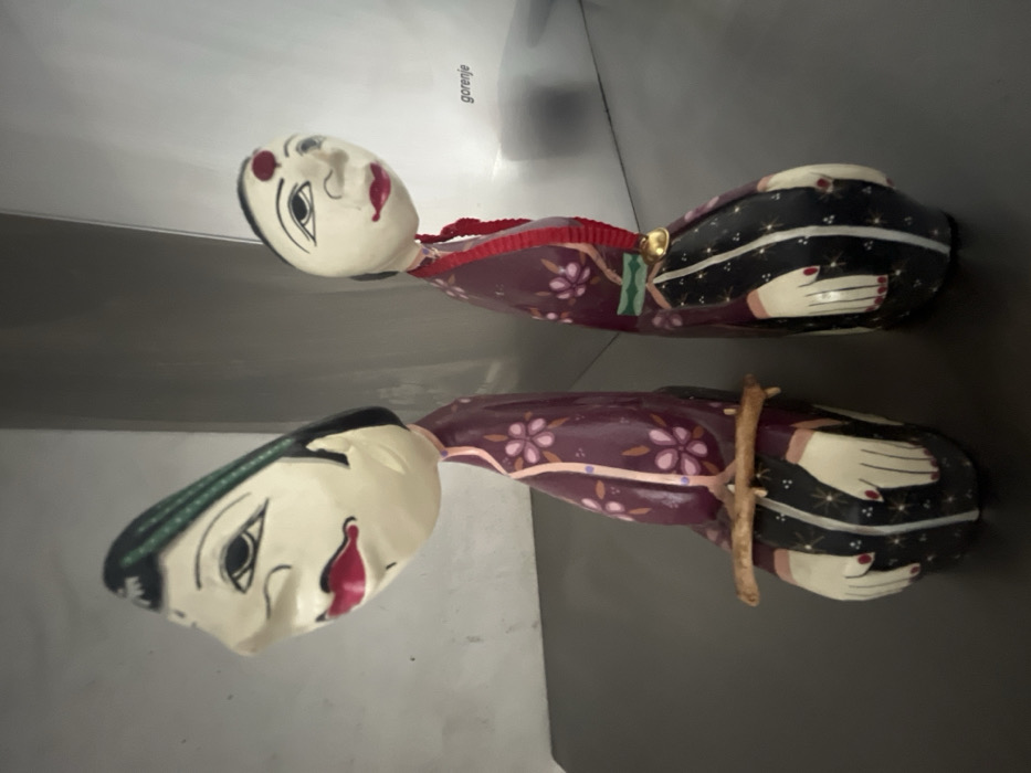 The scene, located in Budva, Montenegro, features two stylized carved wooden figures displayed side-by-side on a flat, dark grey surface against a light grey background. Both figures possess pale, elongated faces with black hair, defined black eyebrows, black eyes, and red lips. The figure on the left has dark green accents in its hair and holds a thin, light-brown stick horizontally across its midsection. It is dressed in a dark purple garment with pink and white floral patterns, transitioning to a black lower section detailed with white and gold linear and star-like motifs, and stylized painted hands. The figure on the right exhibits a red bindi on its forehead and wears a similar dark purple floral-patterned top. A red braided cord, adorned with a small gold bell, hangs from its neck, extending over a light green horizontal stripe and matching black and gold patterned lower garment. The word "gorenje" is subtly visible on the upper right portion of the background, indicative of a household appliance or fixture.