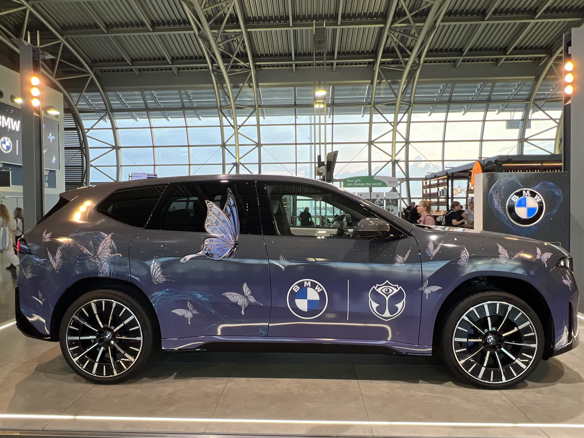 The image shows a BMW iX electric SUV on display indoors, likely at an airport or exhibition hall, given the large glass windows and exposed industrial ceiling structure. The car is adorned with a custom "Tomorrowland" themed wrap featuring numerous butterflies in various sizes and shades of blue and white, set against a dark, starry backdrop. Two logos are visible on the side of the car: the BMW circular logo and the distinctive "Tomorrowland" eye logo.
In the background, several people are milling around, some standing and others walking. The environment appears to be a public space with a modern aesthetic, characterized by metal beams, ribbed ceiling panels, and large, grid-like windows that reveal a bright, overcast sky outside. To the left of the car, a tall display with three illuminated amber lights and the "BMW" logo is visible. To the right, a large display featuring the BMW logo within a nebula-like graphic is present. The floor is a smooth, light-colored surface, with subtle lighting strips embedded in the floor in the foreground.
The specific location is Brussels Airport in Zaventem, Belgium, as indicated by the presence of the "Tomorrowland" branding, a festival that originates in Belgium, and the overall architecture typical of modern airport terminals.
