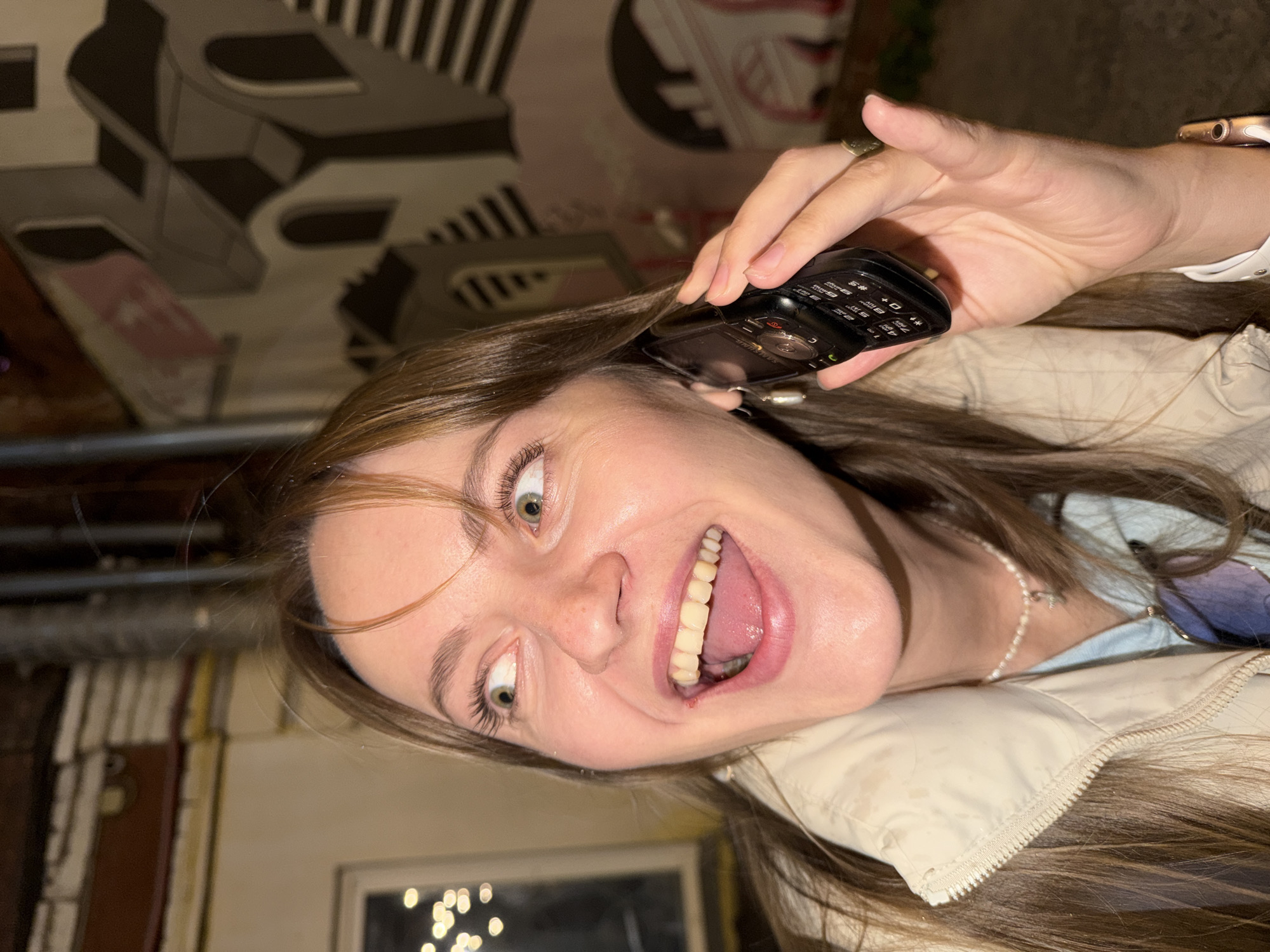 A young woman is holding a black Samsung flip phone to her ear, grinning widely with her mouth open. She is wearing a light-colored jacket, a pearl necklace, and a gold Apple Watch. The background shows a graffitied wall. The location appears to be near a building in Krasnoyarsk, Russia.