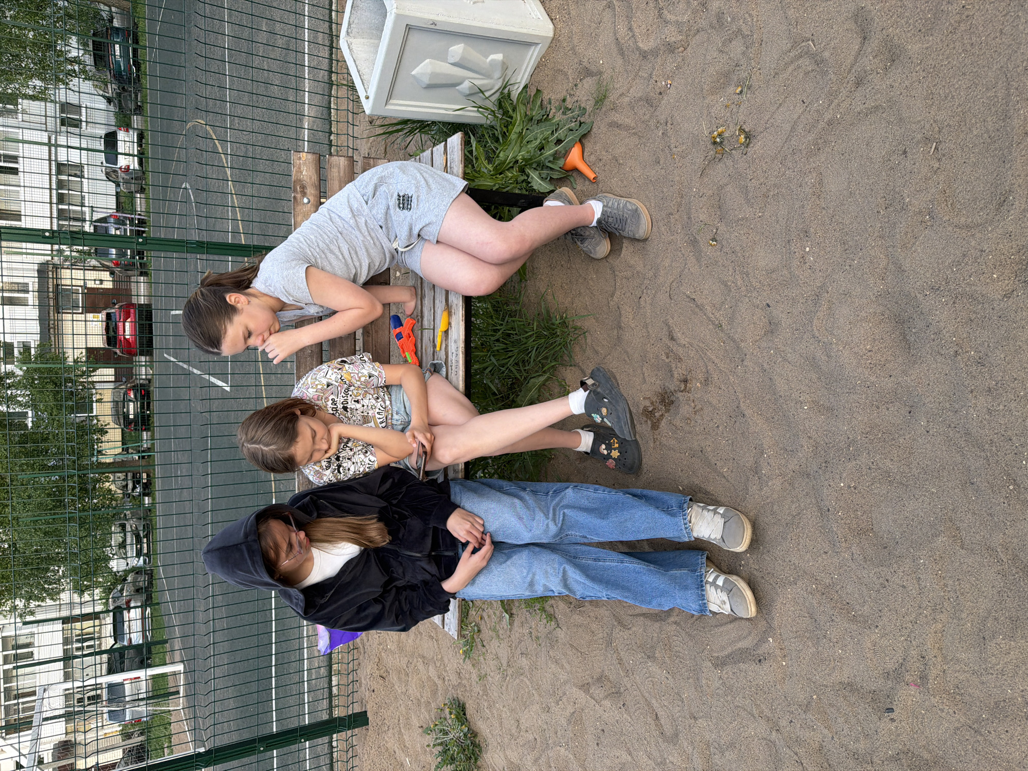 The image depicts three females sitting on a bench. The location is Chaikovsky, Russia. The bench is located next to a sand area and adjacent to a green chain-link fence. Behind the fence, there is a playground area with white lines visible on the ground and a building with multiple windows in the background.
The first female is sitting closest to the top of the image. She is wearing a gray t-shirt and gray shorts, with gray sneakers. The second female is sitting in the middle, wearing a light-colored patterned shirt and jean shorts. The third female is at the bottom, wearing a black hoodie, a white t-shirt, blue jeans, and gray sneakers.