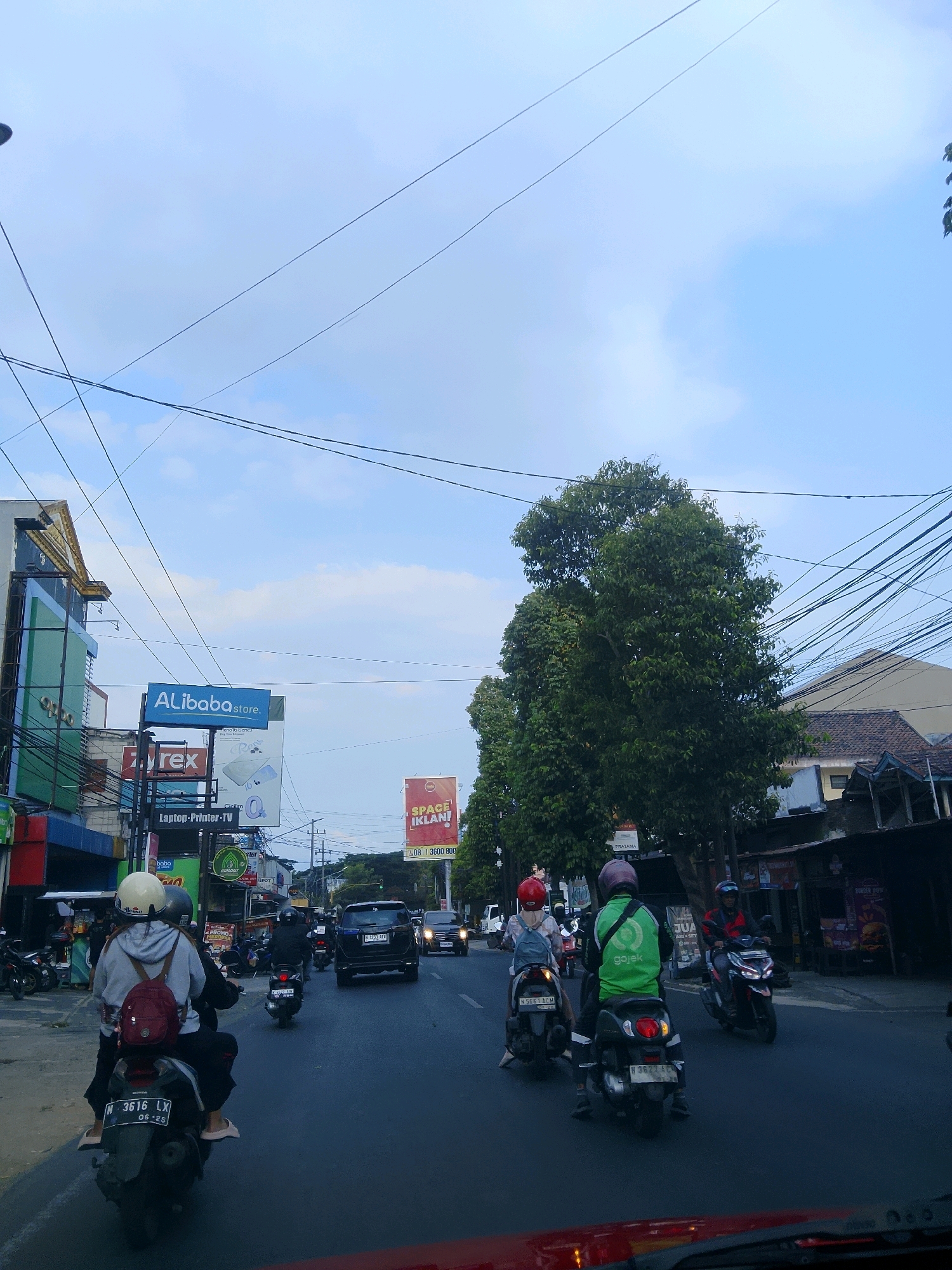 The image shows a street scene in Malang, Indonesia, on a cloudy day. The perspective is from inside a vehicle, looking forward. The road is paved and there are multiple lanes of traffic. Several motorcycles and cars are present, with most of the motorcycles carrying two passengers. On the left side of the road, there are buildings and shops with various signs, including one for "Alibabastore." A large tree with dense foliage is on the right side of the road. Overhead, there are many power lines crisscrossing the sky. The overall impression is a busy urban street with typical Indonesian traffic and infrastructure.