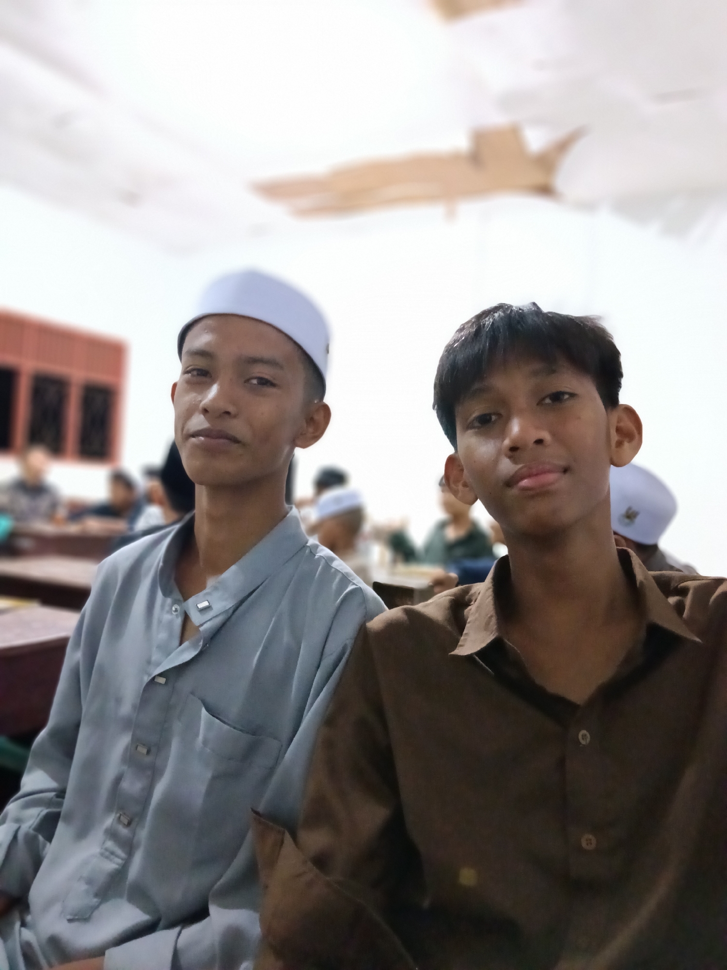 The image shows two young men in what appears to be a classroom or meeting space in Lhokseumawe, Indonesia. The man on the left is wearing a light blue/gray traditional shirt and a white cap. The man on the right is wearing a brown collared shirt. Both are looking towards the camera. In the background, other people are seated at desks. The room is illuminated by overhead lights. There are windows in the background as well. The overall environment is indoors.