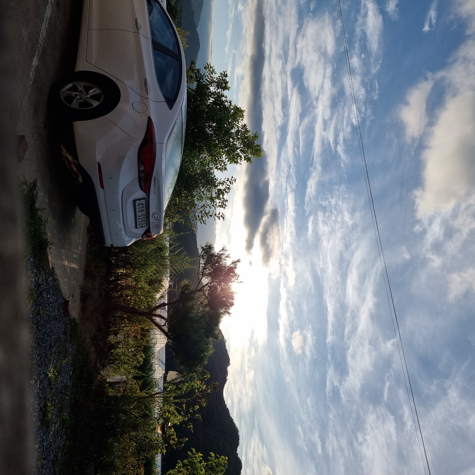 In Dunnae-myeon, South Korea, a white Hyundai Avante sedan is parked in an outdoor lot. The car is facing left, with its rear towards the right side of the frame. Its license plate reads "41라 5250". The car is positioned on a paved surface with white parking lines visible. Behind the car, there is dense green foliage and trees, with a pine tree prominently featured in the mid-ground. In the distance, rolling hills covered in dark green vegetation form the background, under a sky filled with dramatic clouds and illuminated by the setting sun. The sun's rays are breaking through the clouds, creating a bright glare in the upper right quadrant of the image. The scene appears to be a rural or semi-rural area, with greenhouses visible beyond the vegetation in the background. The overall impression is that of a serene, late afternoon or early evening landscape.