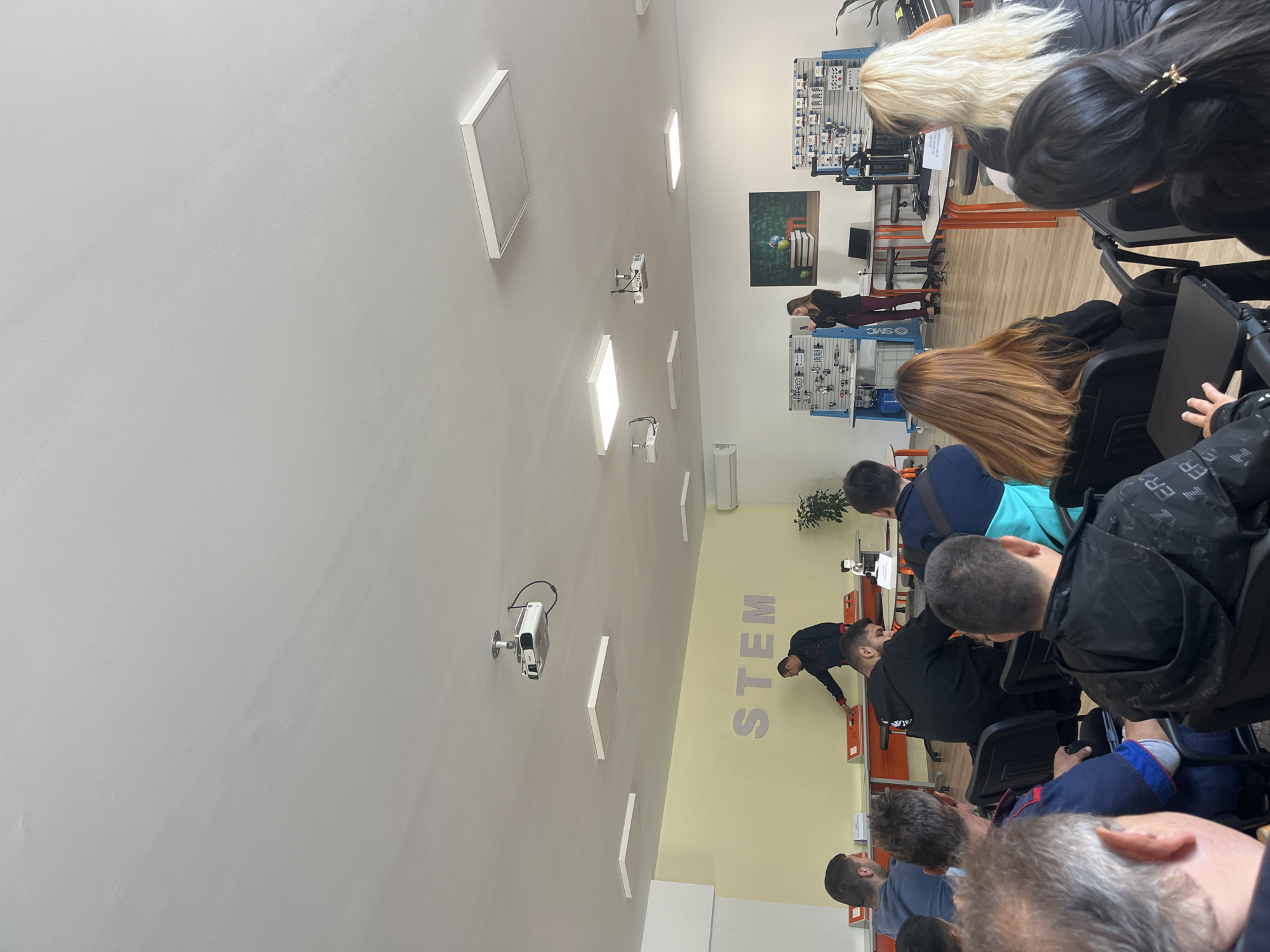 The image shows an indoor scene of a classroom or laboratory setting in Karnobat, Bulgaria. A group of people are seated in rows of chairs, facing a wall with "STEM" written in large letters. There are white rectangular light fixtures and projectors mounted on the ceiling. A woman is standing in the front of the room, interacting with equipment on a table, while others appear to be observing. More equipment is visible along the wall, suggesting a technology or science-focused environment.