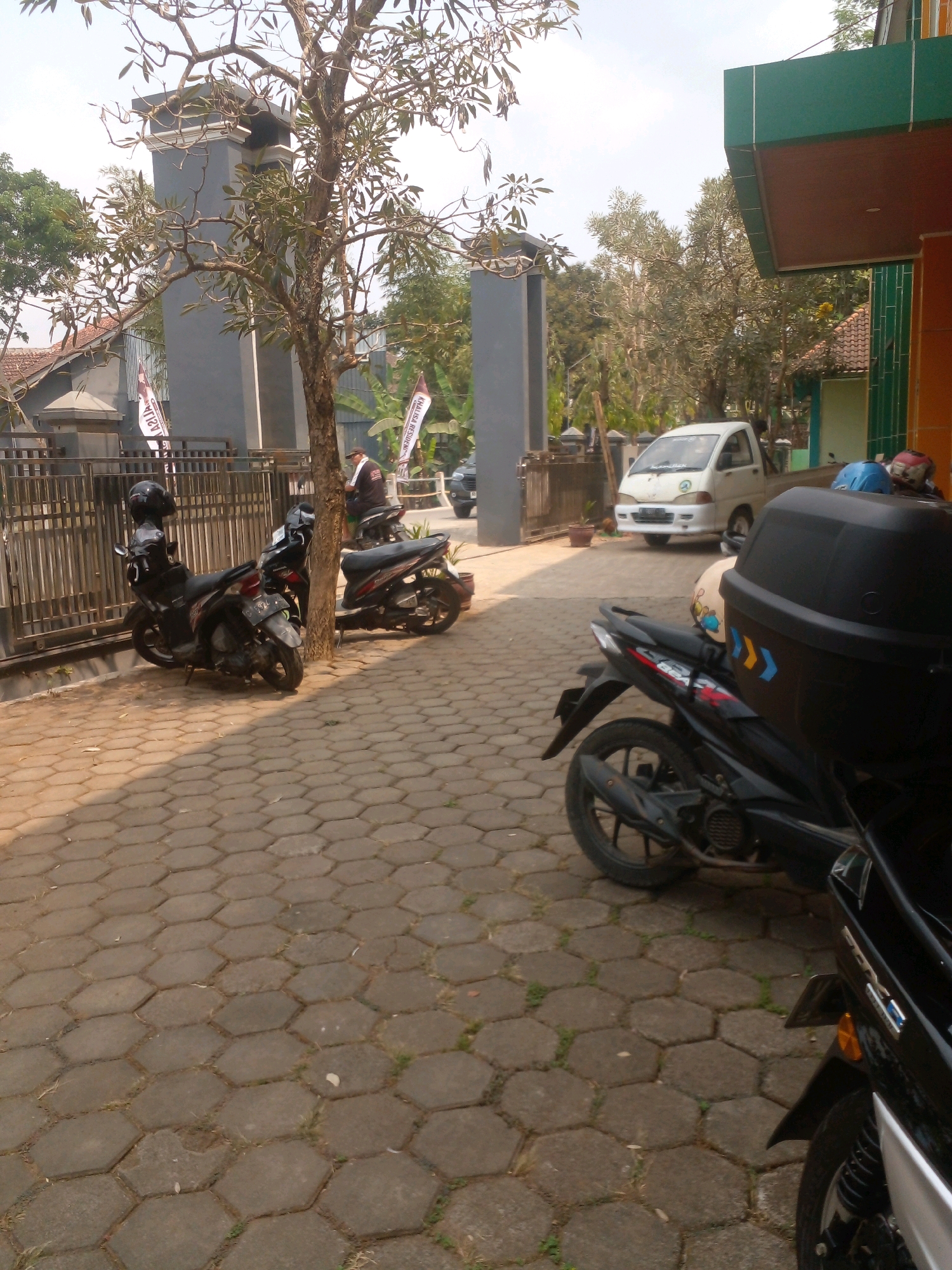In Banyuputih, Indonesia, an outdoor scene is captured with multiple motorcycles parked on a hexagonal paver sidewalk in front of a building with a green and brown facade. A white pickup truck is visible in the background. A mature tree with sparse foliage stands on the right side of the frame, casting a shadow. The environment appears to be a residential or commercial area, with a fence and gate to the left. Two flags, one white with red text and another with white and red elements, are planted near the fence. The sky is overcast.